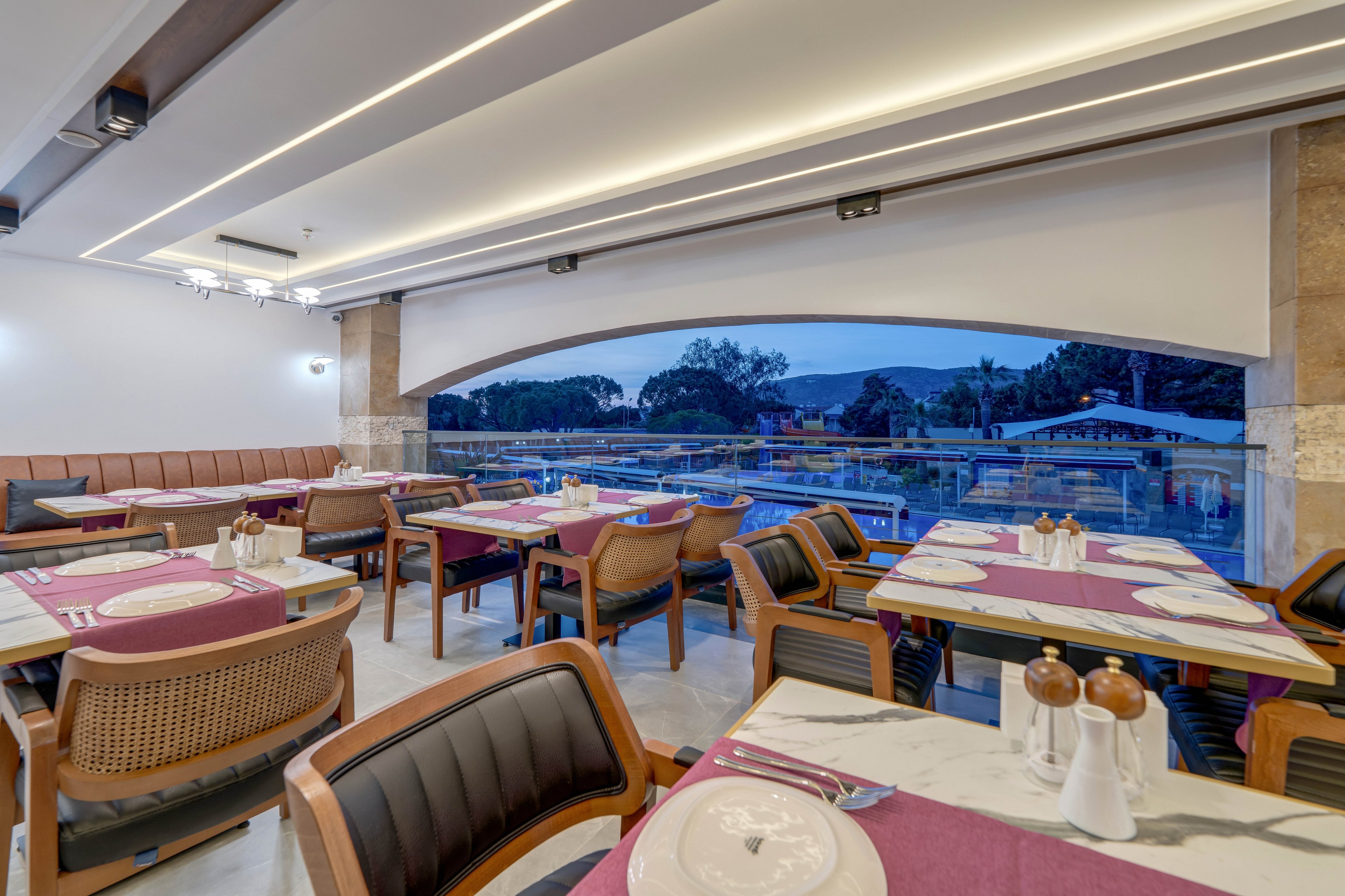 A contemporary restaurant dining area features neatly arranged tables with purple runners and white plates. Wooden chairs with cushioned backs surround marble-topped tables, all set for guests. Large open windows reveal an outdoor pool area and trees at dusk, creating an inviting atmosphere. Soft ceiling lighting and clean architectural lines contribute to the modern, relaxed ambiance.
