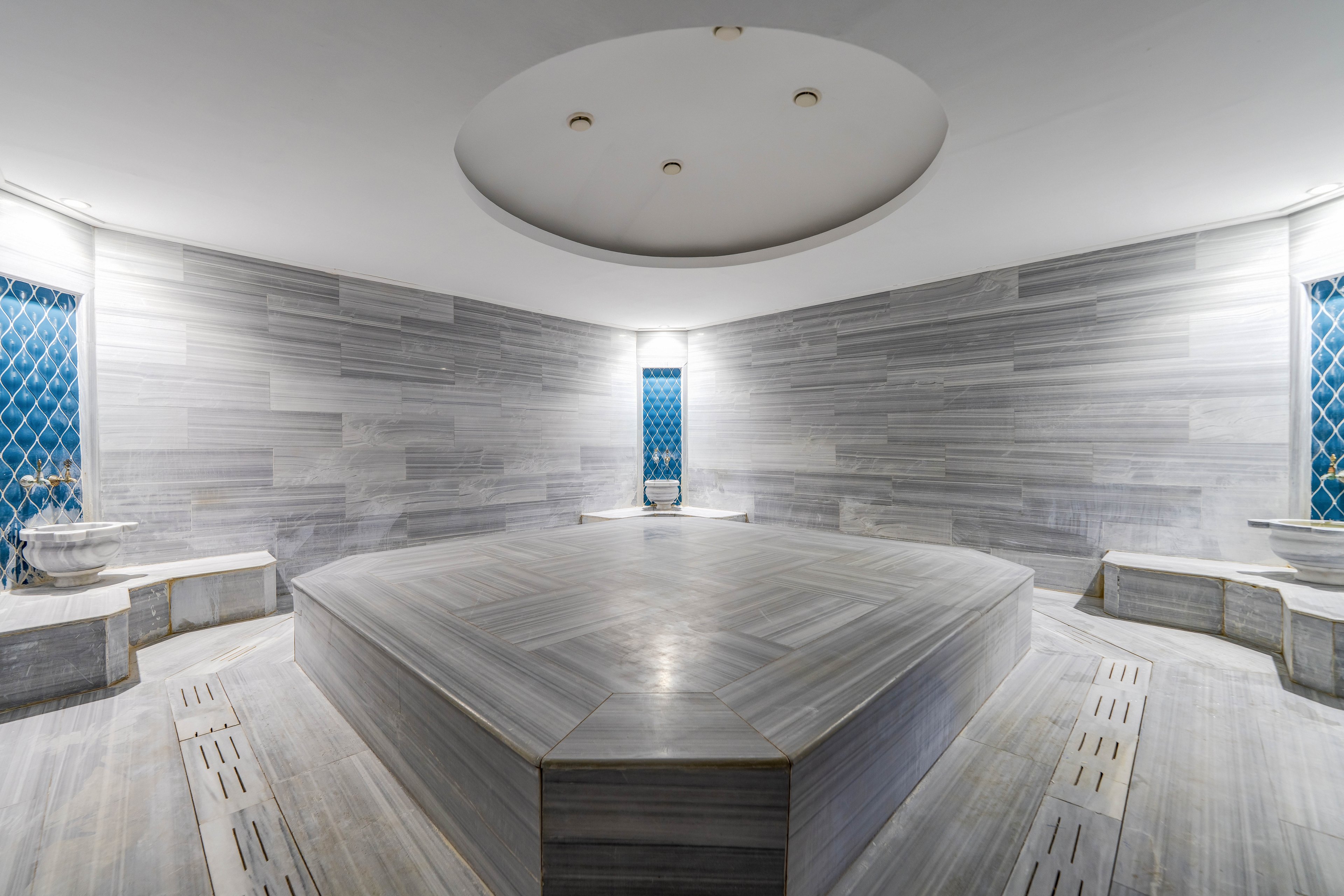 Spacious traditional hammam interior featuring a large central marble platform. The room is lined with smooth gray stone walls and built-in benches along the sides. Decorative blue-tiled windows add color and pattern to the otherwise minimalist space. Overhead ceiling lights softly illuminate the clean, serene bathing environment.