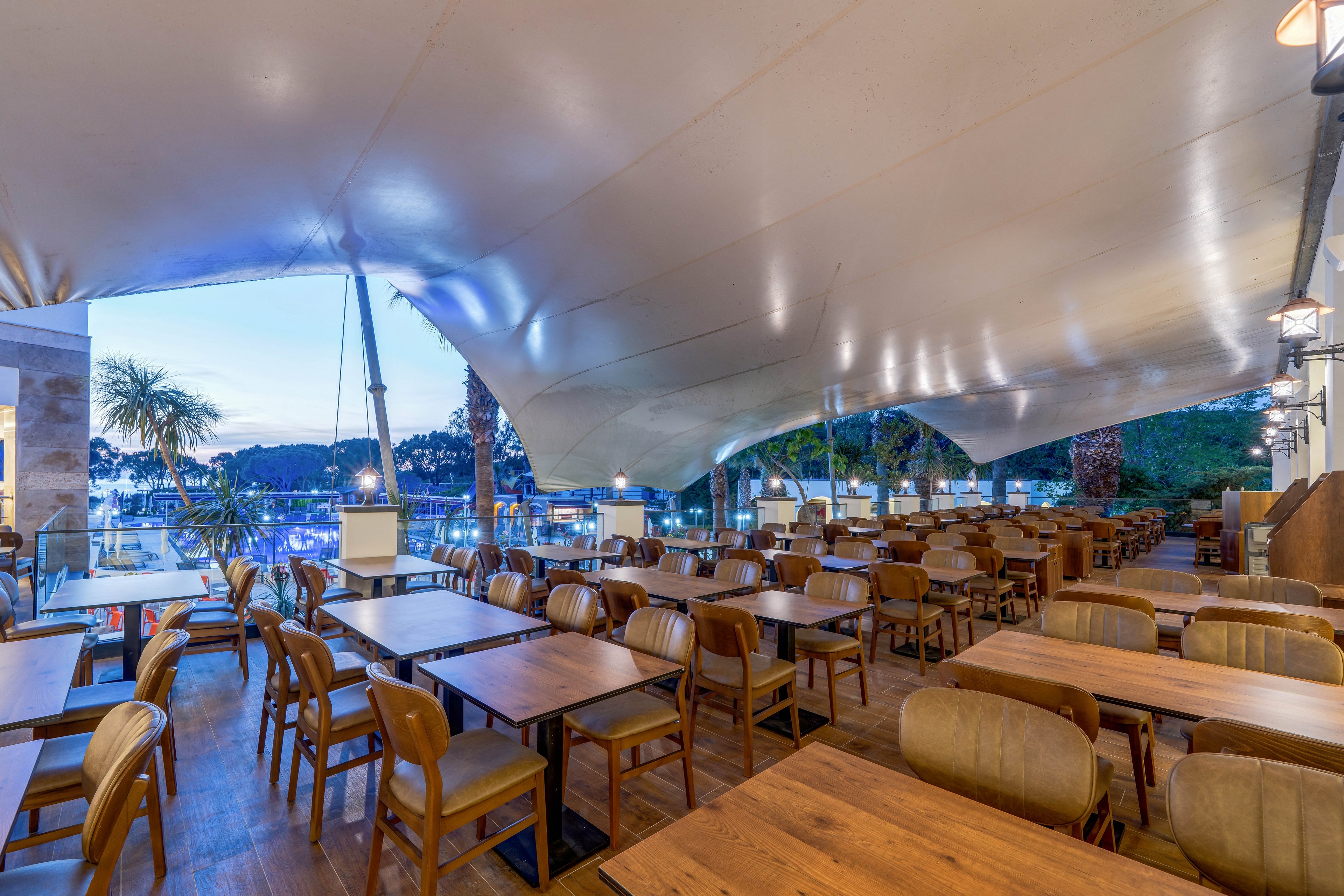 An open-air restaurant terrace features rows of wooden tables and cushioned chairs neatly arranged. A large white canopy stretches overhead, providing shelter while still allowing views of the evening sky. Soft ambient lighting illuminates the seating area, contrasting with the cool blue tones of the twilight outside. In the background, palm trees and distant lights suggest a waterfront or resort setting.