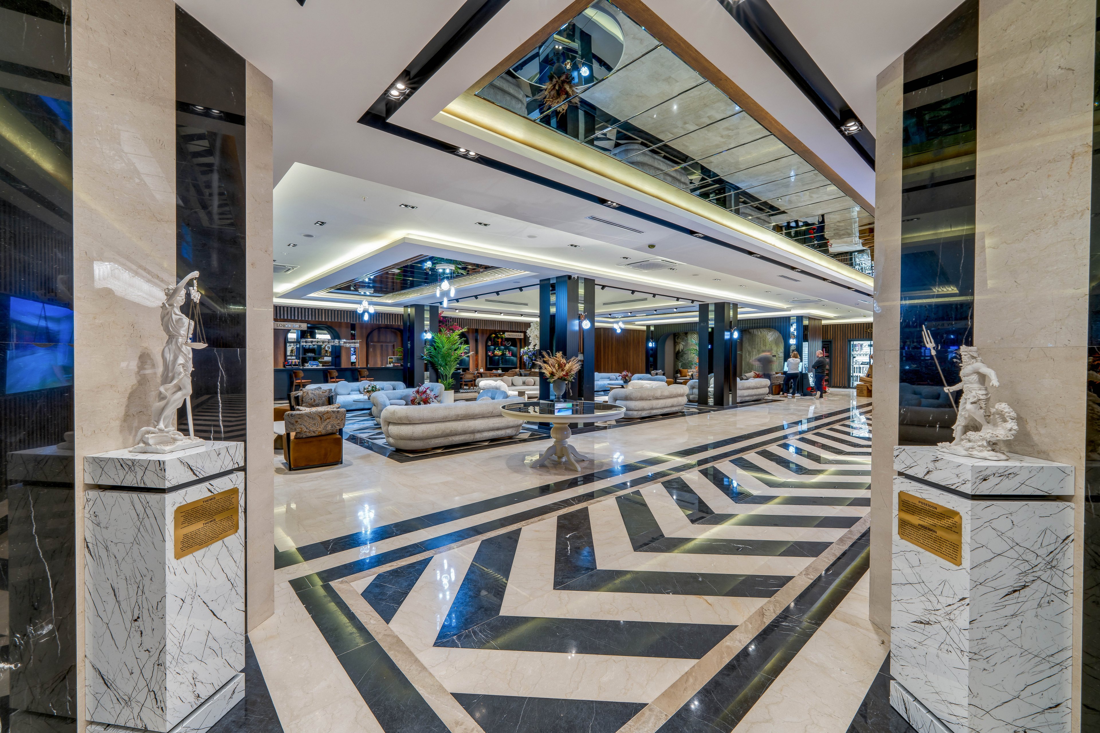 A spacious, luxurious hotel lobby features polished marble floors with bold black and white geometric patterns. Plush sofas and seating areas are arranged in the background, creating an inviting social space. Decorative statues on marble pedestals frame the entrance, while mirrored ceilings and recessed lighting enhance the sense of depth and brightness. The overall design emphasizes modern elegance and high-end hospitality.