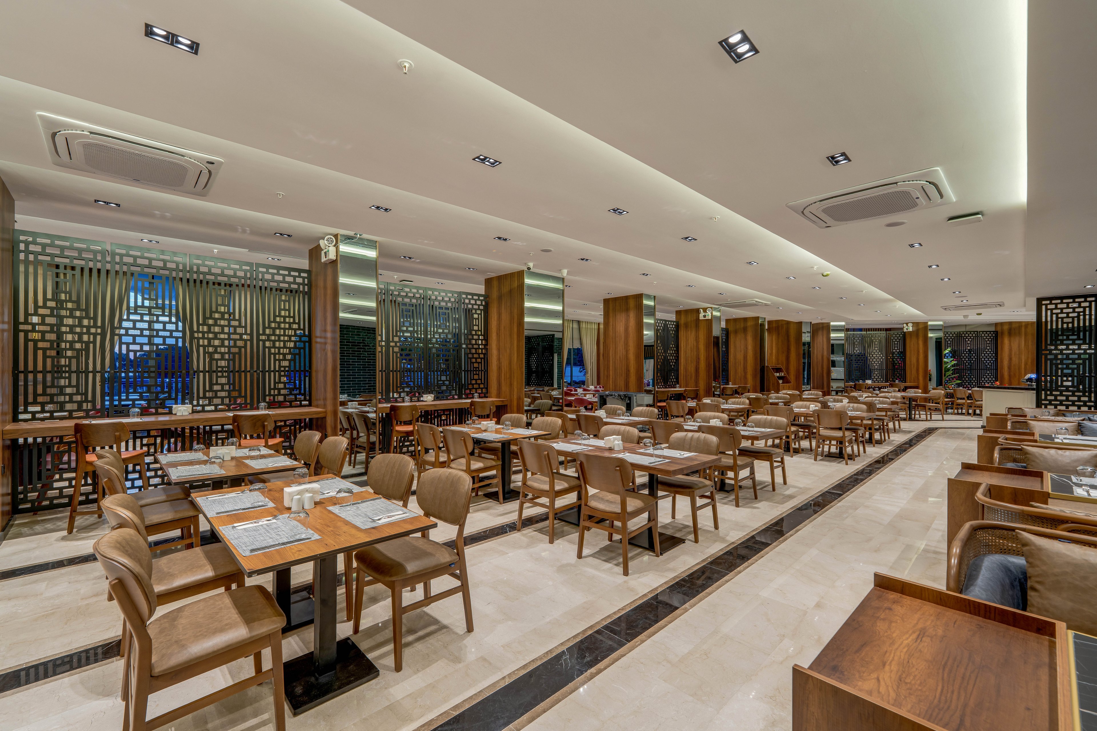 A spacious contemporary restaurant dining area is shown with neatly arranged wooden tables and upholstered chairs. The room is empty of guests, emphasizing the clean, orderly setup with place mats and table settings prepared. Warm wood tones contrast with light stone flooring and a bright, evenly lit ceiling. Decorative partition panels and large windows add depth and a sophisticated, modern ambiance.