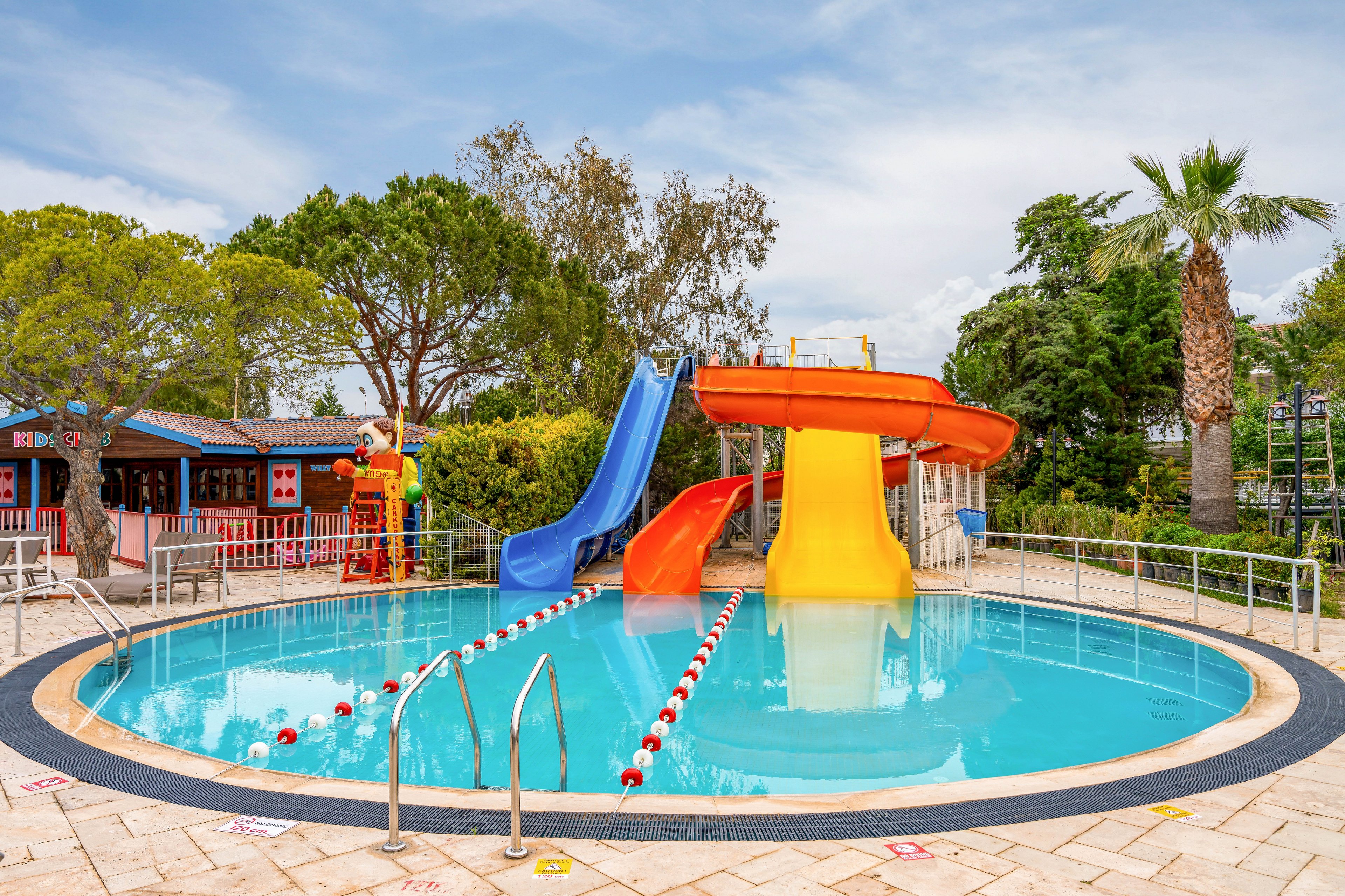 A small circular swimming pool features bright blue, yellow, and orange water slides leading into the clear water. The pool is empty and surrounded by metal handrails and a tiled deck with depth markers. Red and white floating lane dividers stretch across the pool, and a low fence encloses the area. In the background, trees, palms, and resort buildings create a relaxed holiday atmosphere under a partly cloudy sky.