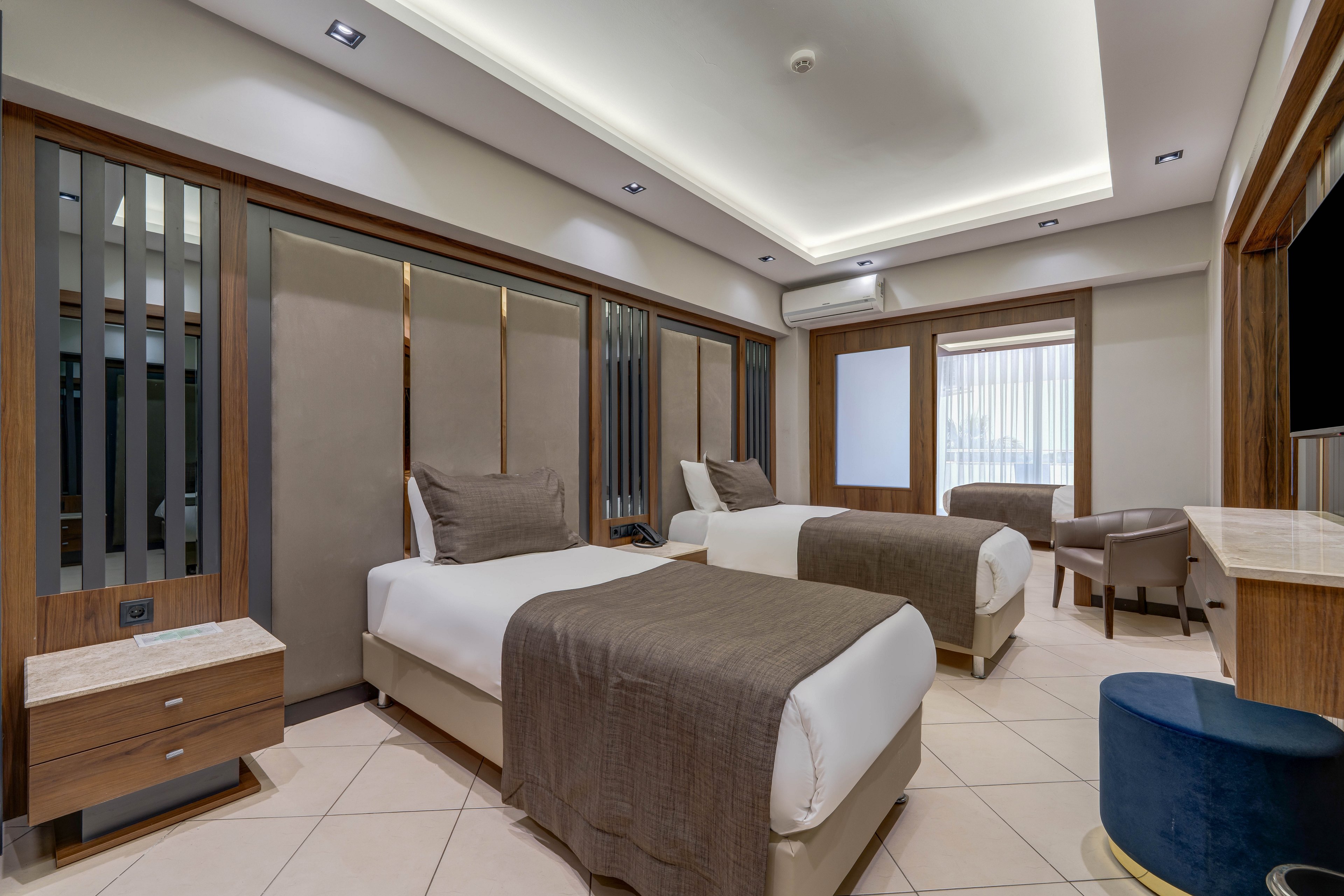 Contemporary hotel room featuring two neatly made twin beds with brown throws and pillows. The space includes wood-paneled walls, built-in headboards, and a recessed ceiling with soft lighting. A small bedside table, upholstered armchairs, and a wall-mounted television complete the comfortable setting. Neutral tones and clean lines create a calm, minimalist atmosphere suitable for business or leisure stays.
