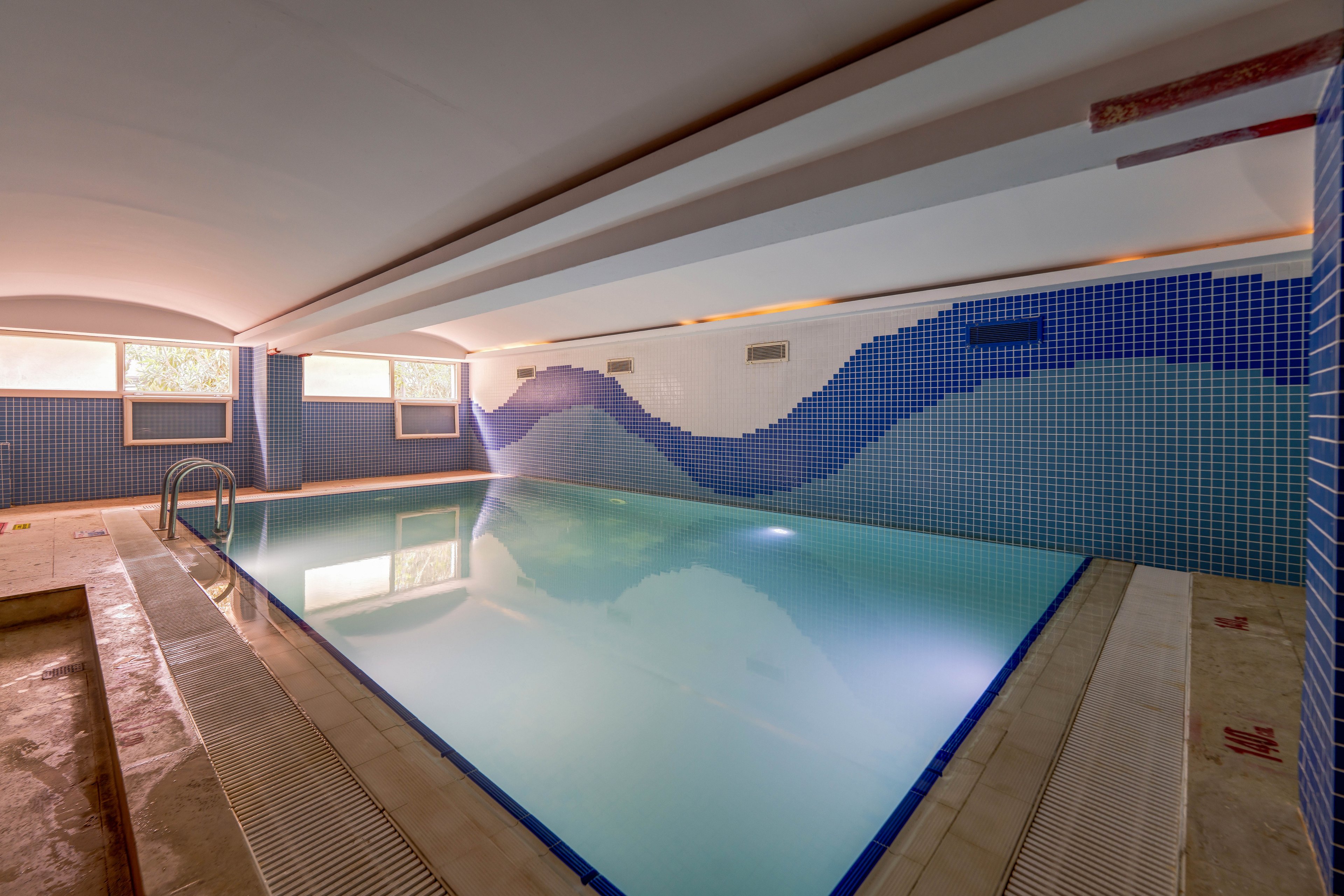 An empty indoor swimming pool is shown in a modern facility. The pool is surrounded by blue mosaic tiles forming a wave pattern along the walls. Soft natural light enters through rectangular windows, reflecting on the calm water surface. The space appears clean and quiet, with no people present and a serene atmosphere.