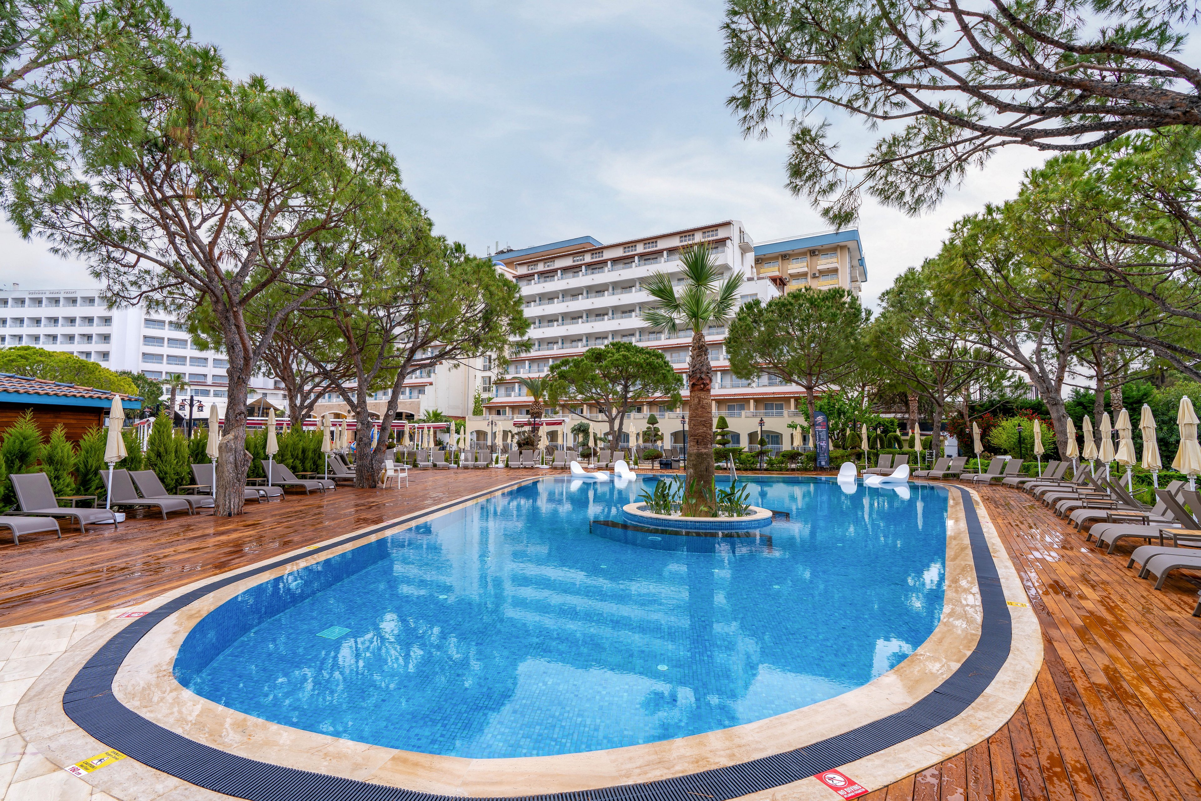An outdoor swimming pool is surrounded by wooden decking and sun loungers at a resort hotel. Tall trees and landscaped greenery frame the pool area, creating a relaxing vacation atmosphere. Multi-story hotel buildings rise in the background under a cloudy sky. Closed umbrellas and empty chairs suggest an off-peak or early morning setting.