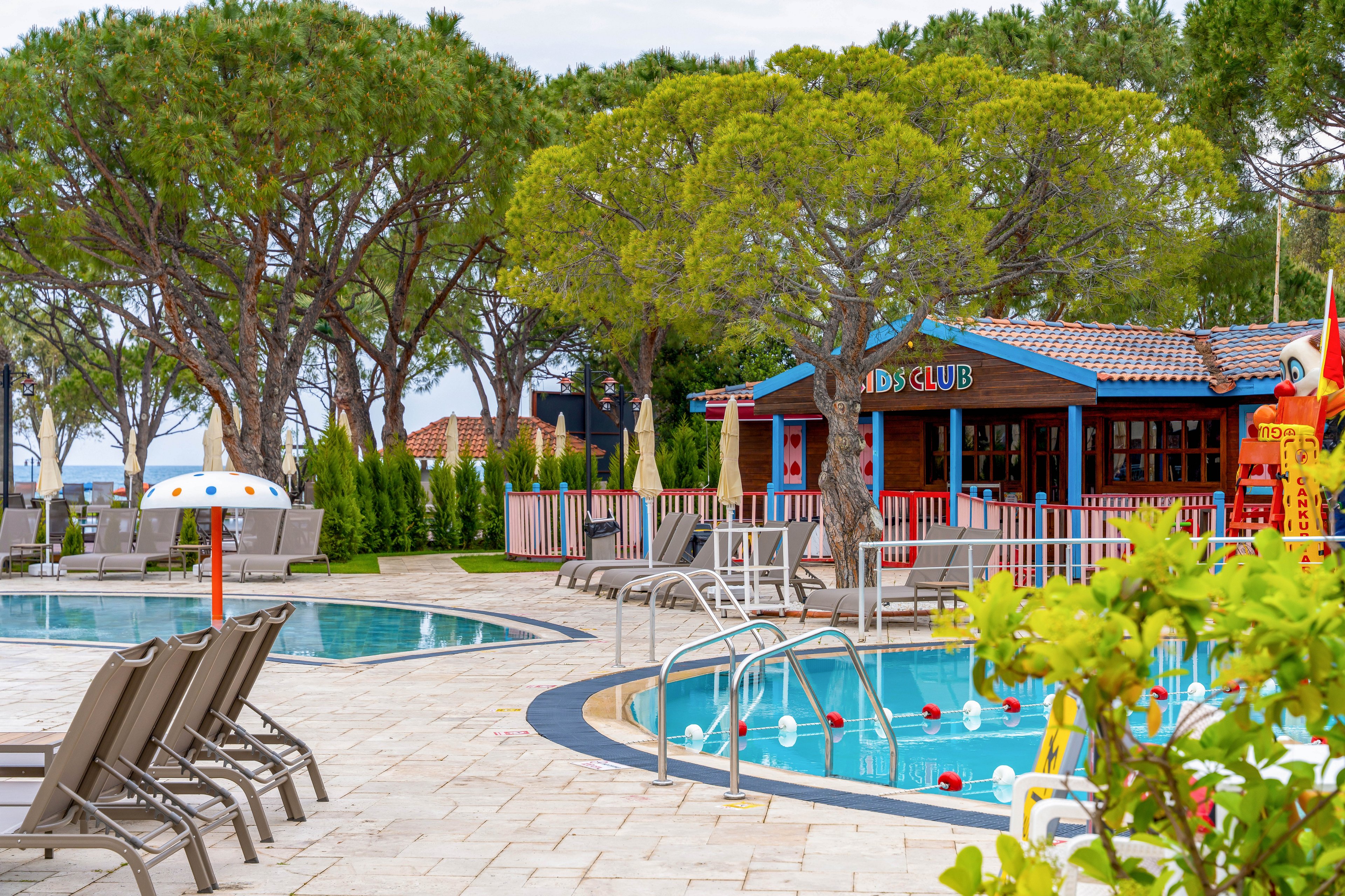 An outdoor resort swimming pool area is surrounded by lounge chairs and tall green trees. A colorful kids club building stands near the pool, featuring bright accents and playful design. The pool has metal ladders, safety ropes with buoys, and a shallow section suitable for children. The overall scene suggests a family-friendly vacation spot with leisure and relaxation by the water.