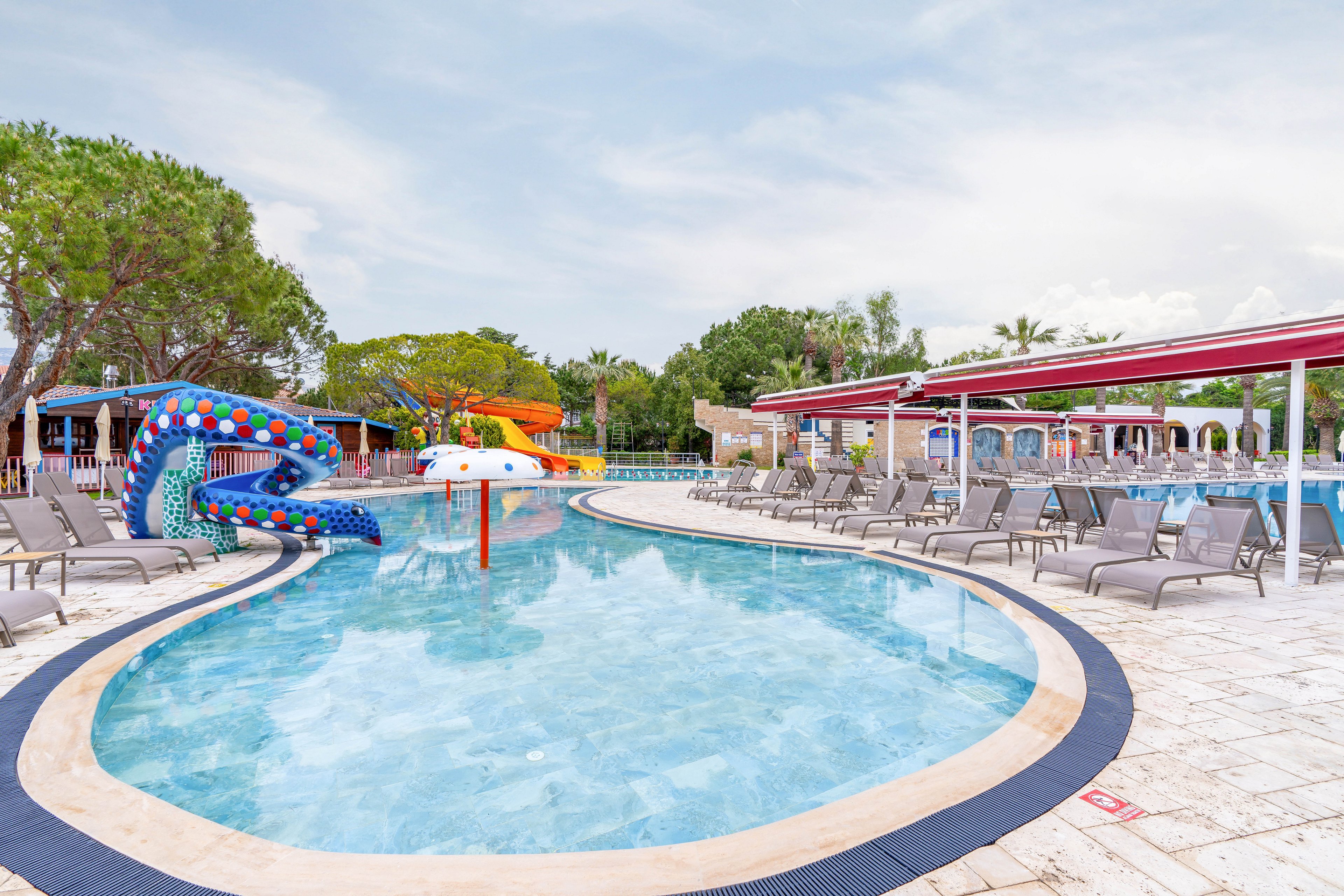 An outdoor resort swimming pool curves through a tiled deck lined with empty lounge chairs. Colorful water slides and playful structures, including a blue serpent-like slide and orange and white slides, stand at the far end of the pool. Tall trees and low resort buildings frame the scene under a bright, lightly clouded sky. The calm water and absence of people create a peaceful, off-peak vacation atmosphere.