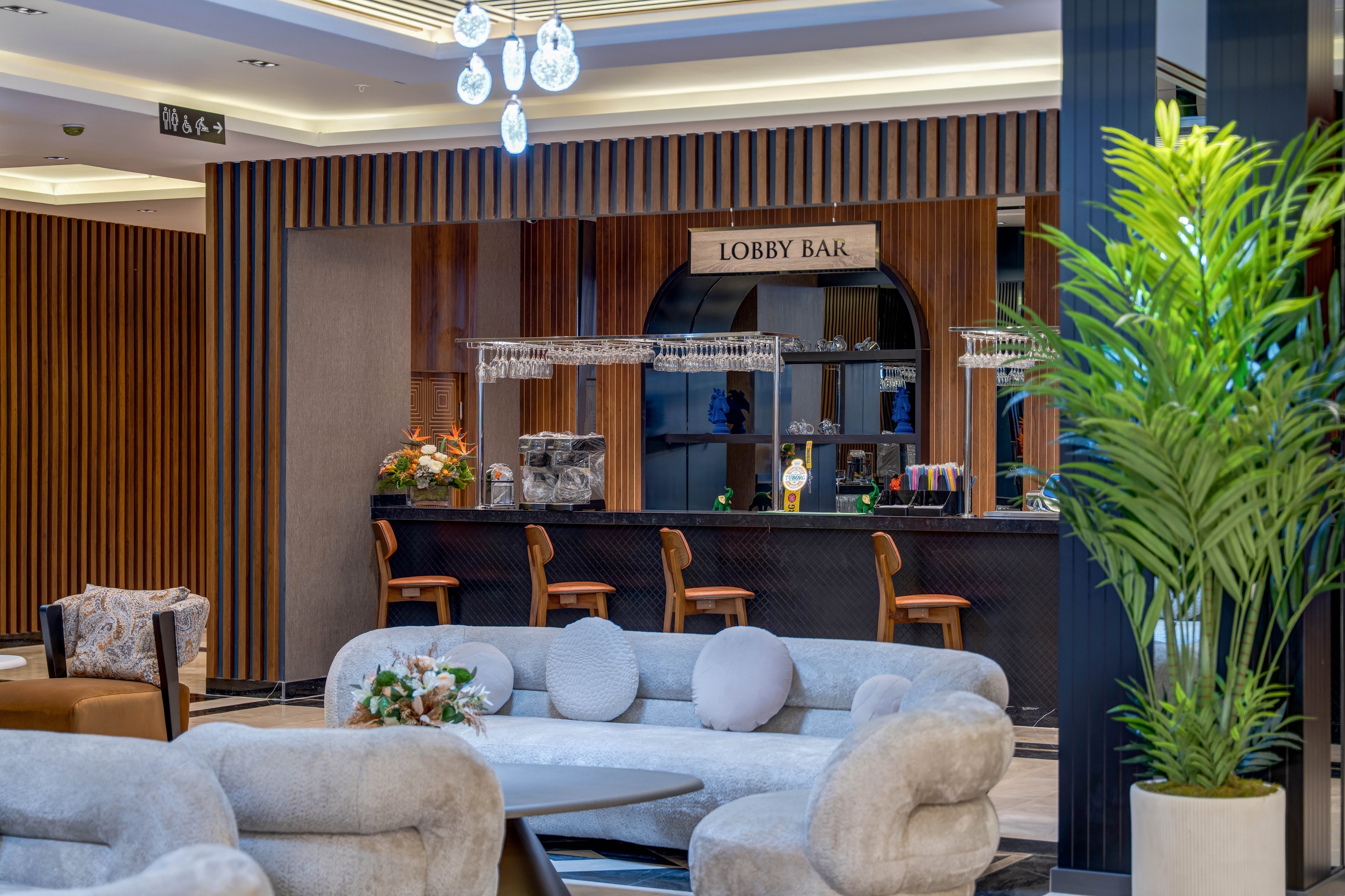 A stylish hotel lobby bar features a sleek counter with several bar stools neatly arranged in front. Soft, plush sofas and armchairs create a comfortable seating area in the foreground, centered around a low round table with a floral arrangement. Vertical wooden slats and dark accents give the space a contemporary, upscale look. Warm lighting and decorative hanging fixtures contribute to a relaxed, inviting atmosphere.