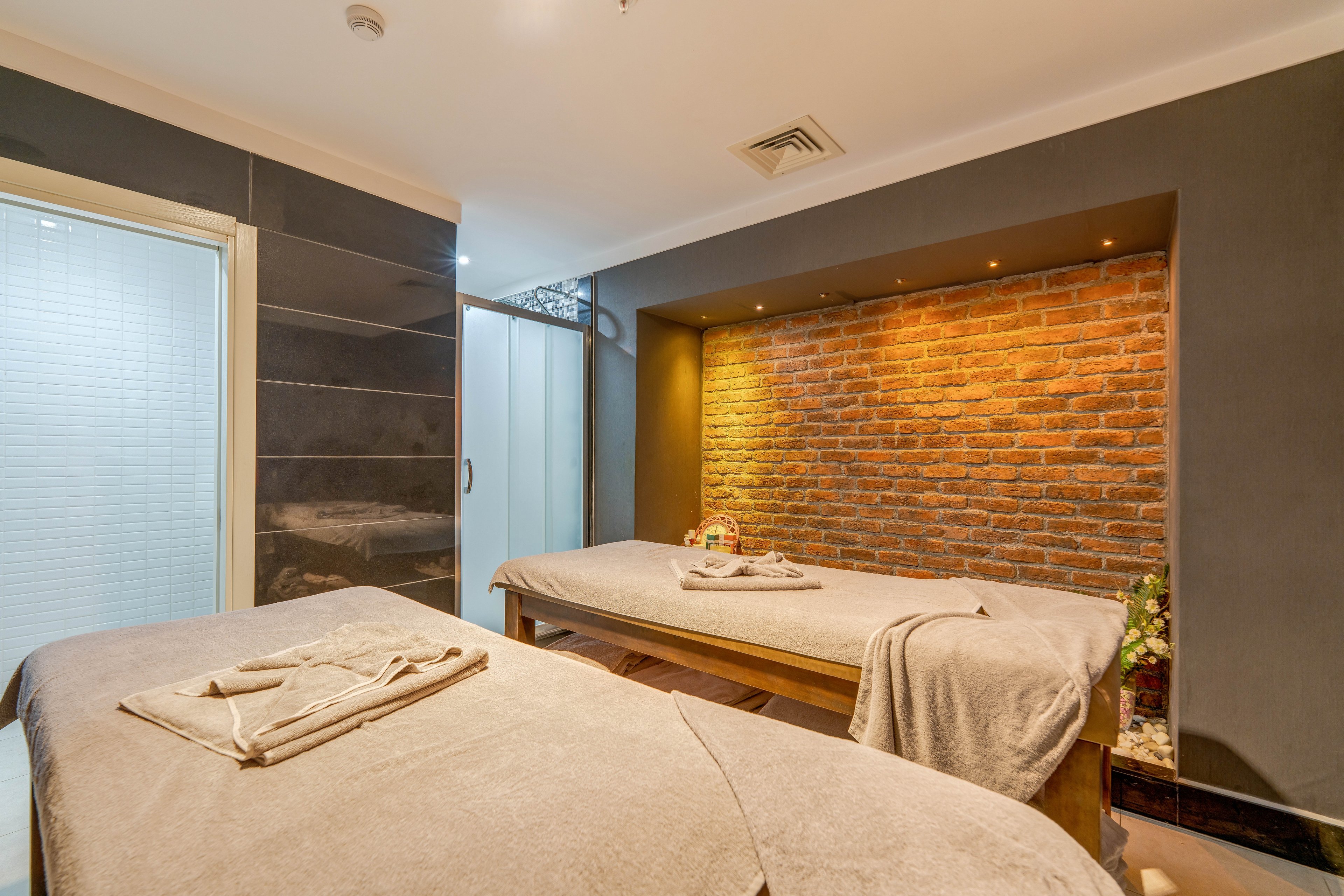 A warm, inviting spa treatment room features two massage tables covered in soft beige linens. The space is accented by a brick feature wall with recessed lighting that creates a relaxing ambiance. Towels are neatly folded on the tables, suggesting readiness for massage or wellness treatments. Modern finishes and soft, indirect lighting contribute to a calm, luxurious atmosphere.