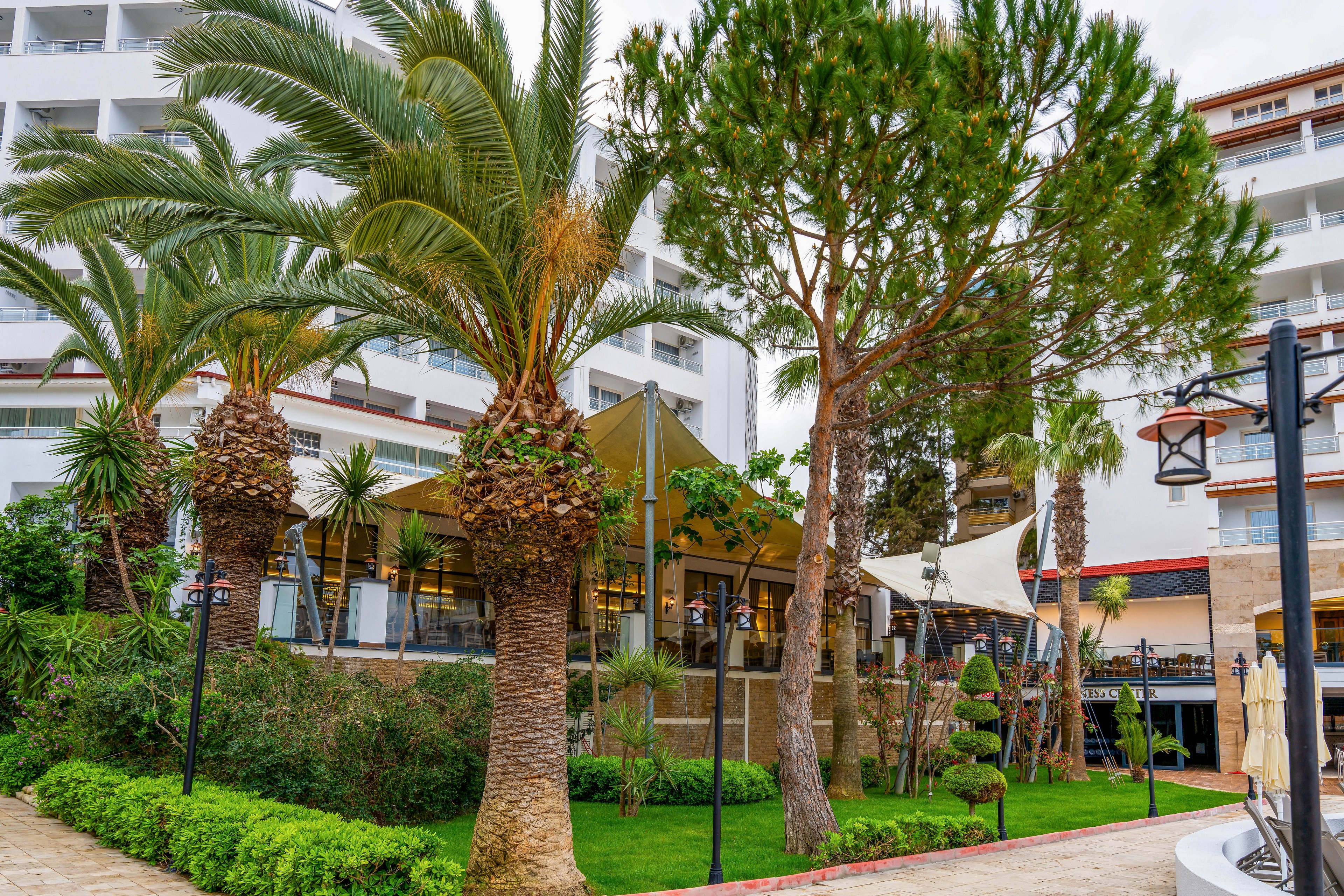 A lush hotel garden features tall palm trees and manicured greenery in the foreground. Behind the trees, a modern multi-story resort building with balconies rises above the landscaped area. Outdoor seating and shaded canopy structures suggest a relaxed dining or lounge space. The scene conveys a vacation atmosphere with well-maintained paths and decorative lighting.