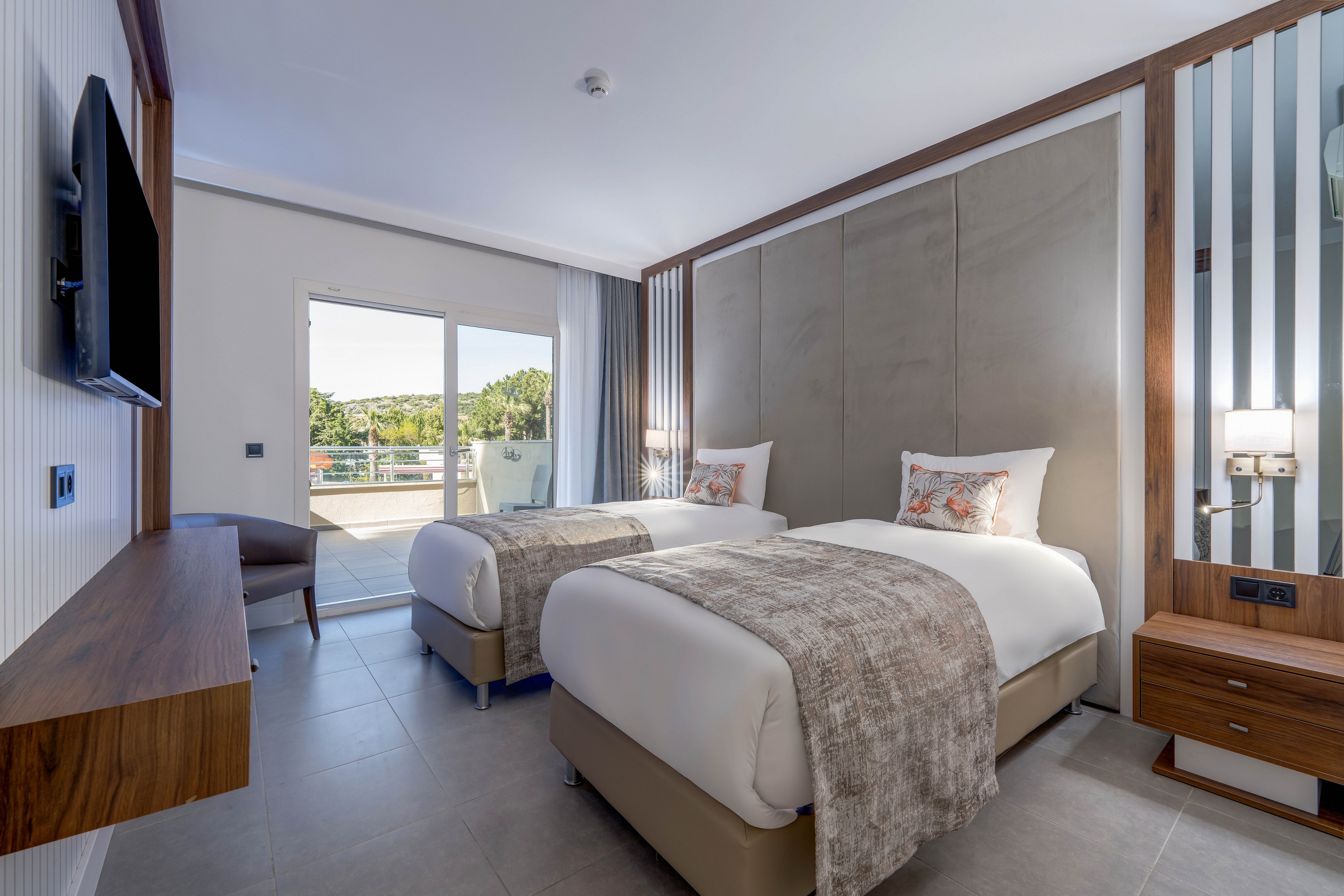 Contemporary hotel room featuring two neatly made twin beds with neutral bedding and accent pillows. The space includes a wall-mounted flat-screen TV, wooden furniture, and a bedside lamp for ambient lighting. Large sliding glass doors open to a balcony with outdoor seating and a view of greenery. The overall design is minimalist with clean lines, soft tones, and ample natural light.
