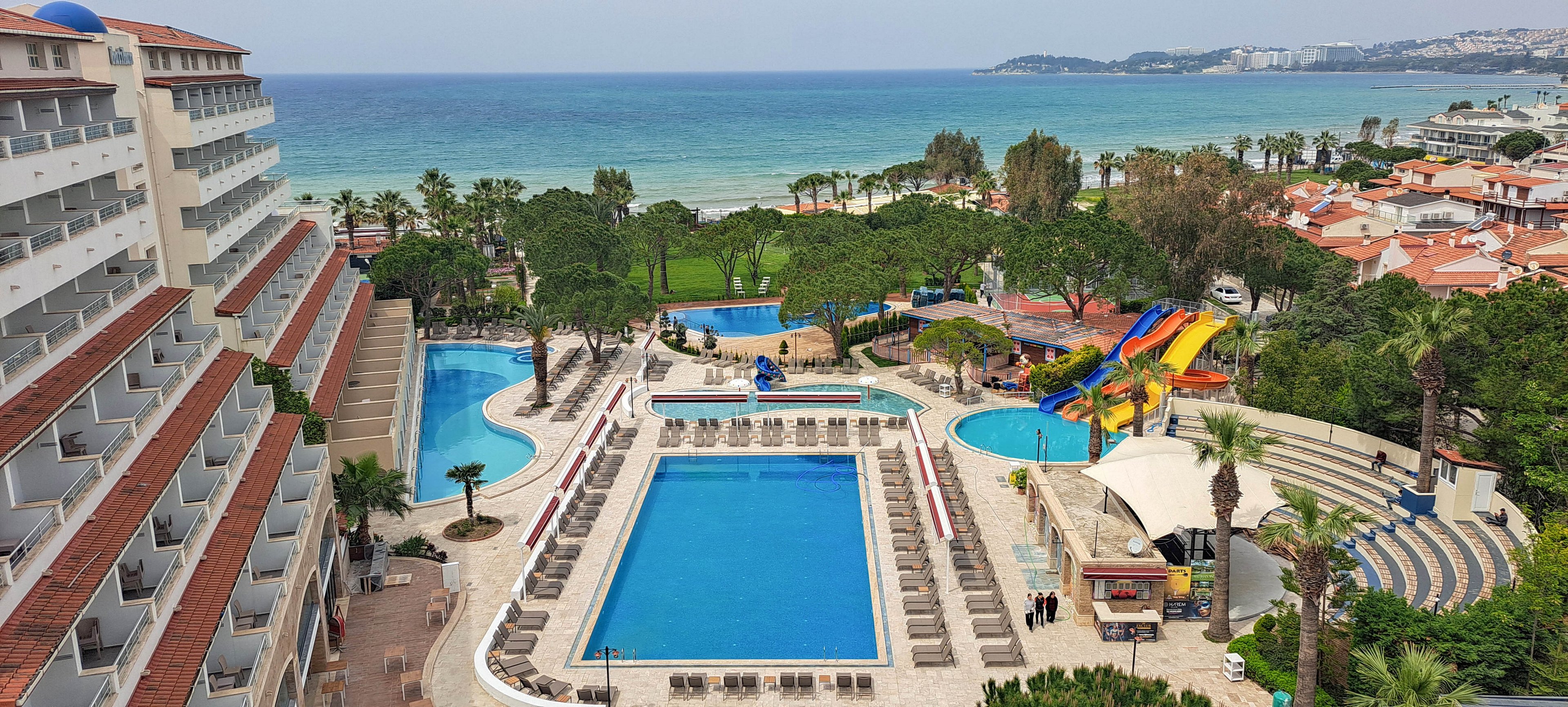 A large seaside resort features multiple outdoor swimming pools surrounded by sun loungers. Colorful water slides and a small water park area are visible near the center, adding a playful attraction for guests. An amphitheater-style seating area and landscaped gardens with palm trees enhance the vacation atmosphere. In the background, the coastline and calm sea stretch toward distant headlands and coastal buildings.