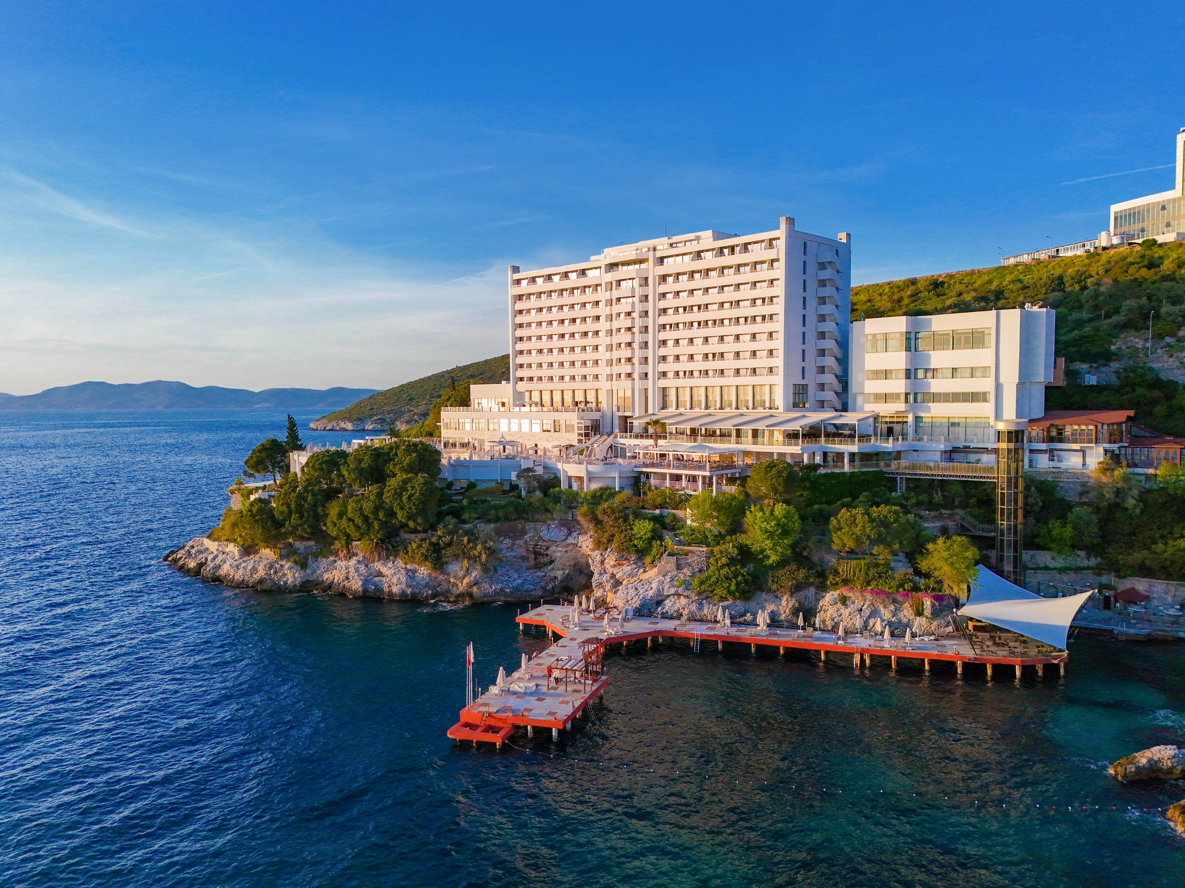A large modern hotel complex stands on a rocky coastline overlooking a calm blue sea. The building rises several stories high with balconies facing the water and surrounding green hills. A red wooden pier extends from the shore into the sea, featuring sunbathing areas and a white canopy structure. The scene is captured in warm daylight, highlighting the clear sky and tranquil coastal setting.