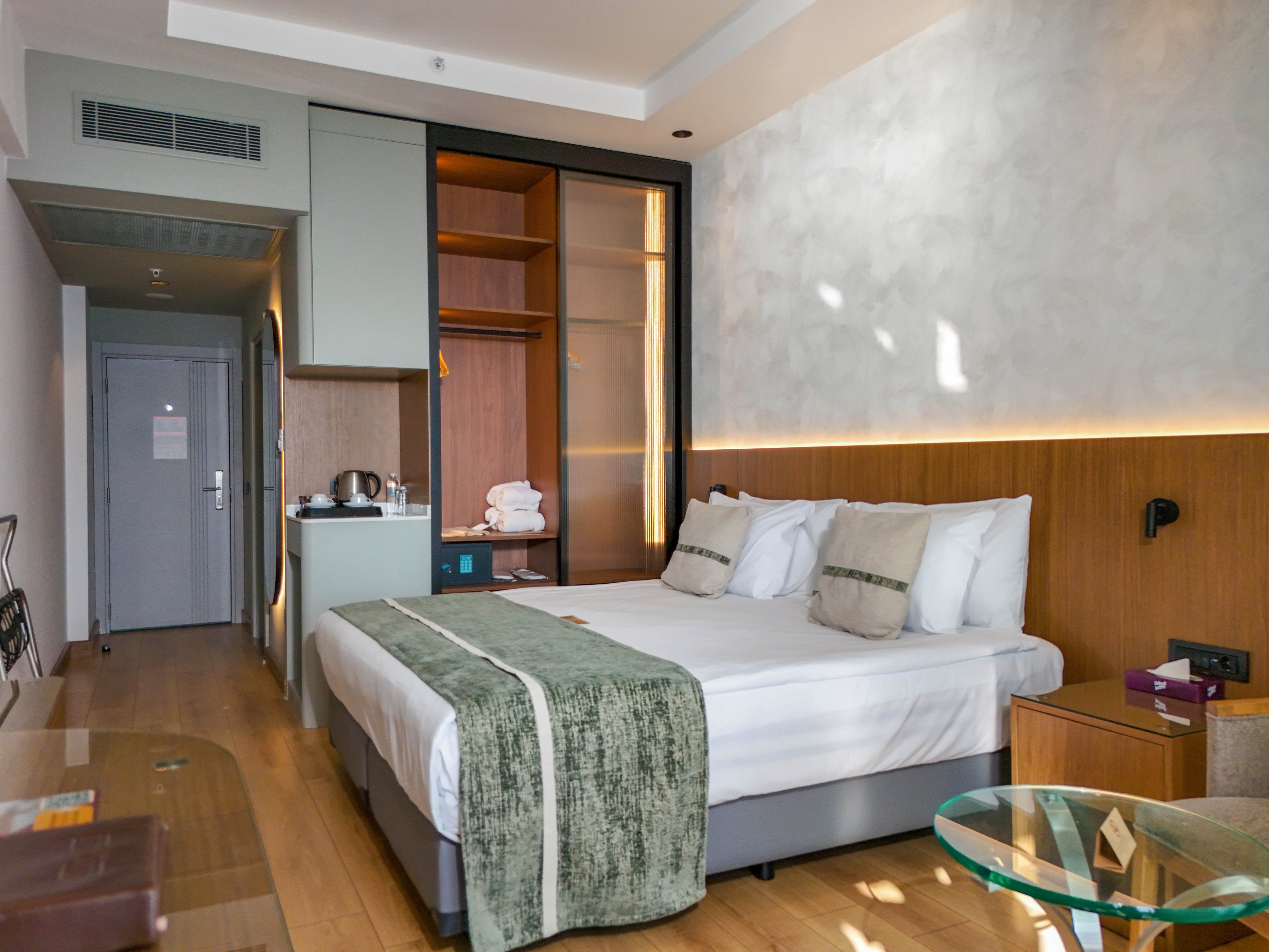 A contemporary hotel room features a neatly made double bed with white linens and green decorative runners and cushions. The space includes built-in wardrobes, a small minibar area with a kettle, and folded towels on a shelf. Warm wooden flooring and paneling contrast with light-colored walls and subtle indirect lighting along the headboard. A glass-topped table and a view toward the entrance door complete the clean, minimalist interior.