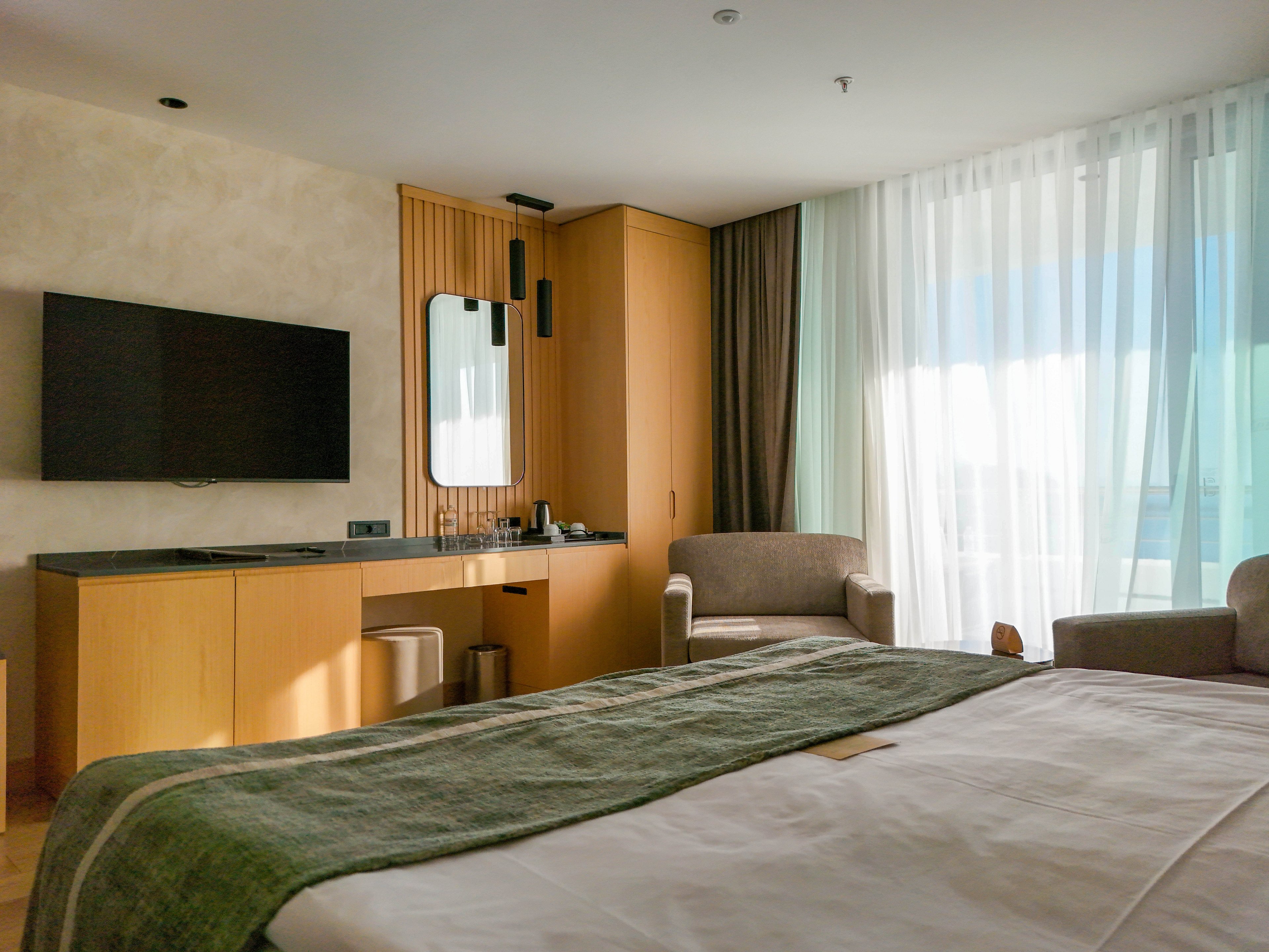 A contemporary hotel room features a neatly made bed in the foreground with a green runner. A wall-mounted flat-screen television and a wooden console with a mirror and lighting fixtures sit along one wall. An upholstered armchair and floor-to-ceiling sheer curtains allow natural light to fill the space. The overall design is minimalist with warm wood tones and soft neutral colors, creating a calm and inviting atmosphere.