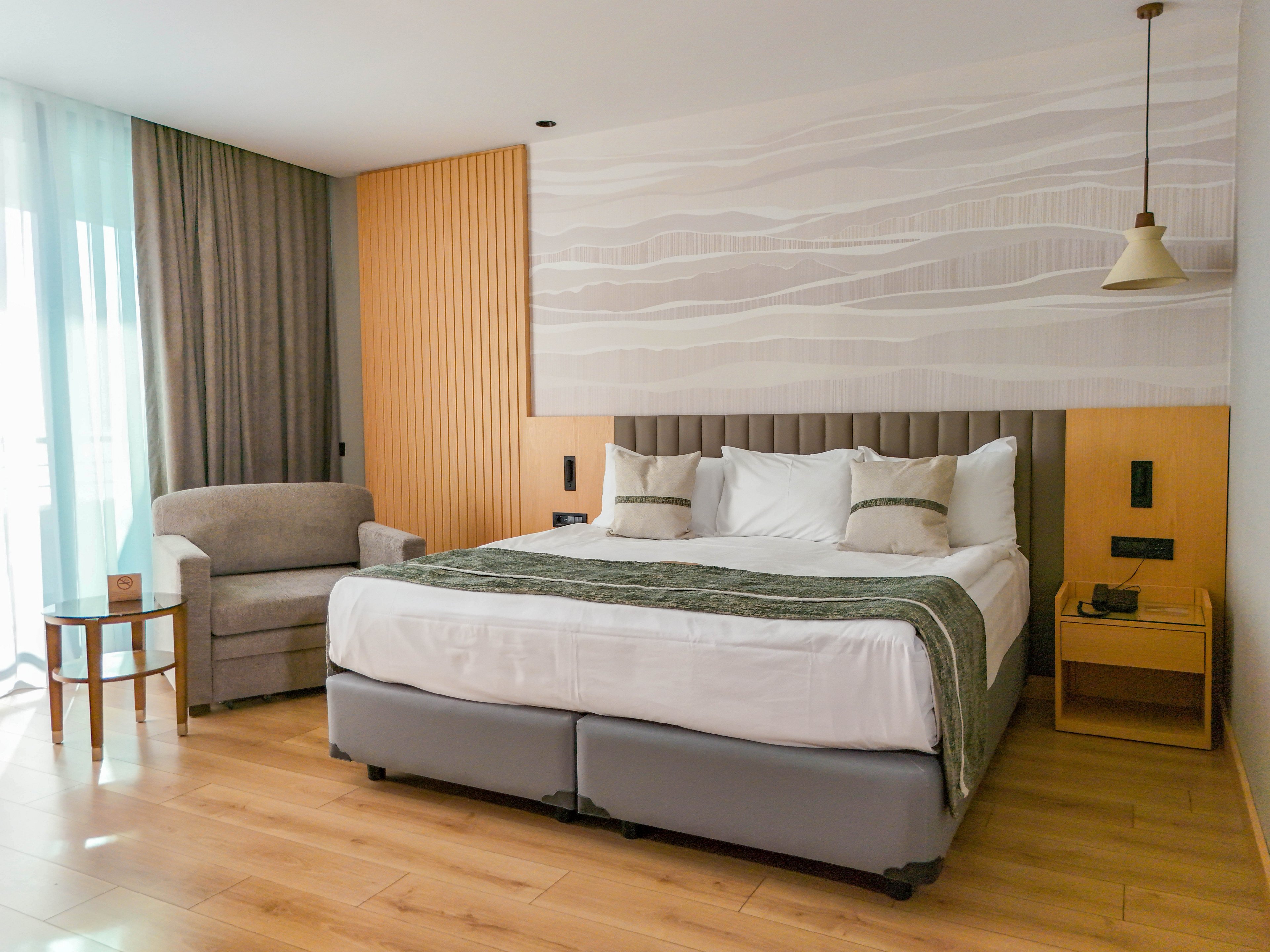 A contemporary hotel bedroom features a large king-size bed with white linens and neutral accent pillows. The room includes a gray upholstered armchair beside a small round side table near a window with sheer and blackout curtains. Warm wooden flooring and paneling complement the minimalist wall art behind the bed. Soft, natural light and simple bedside furniture create a calm, inviting atmosphere.