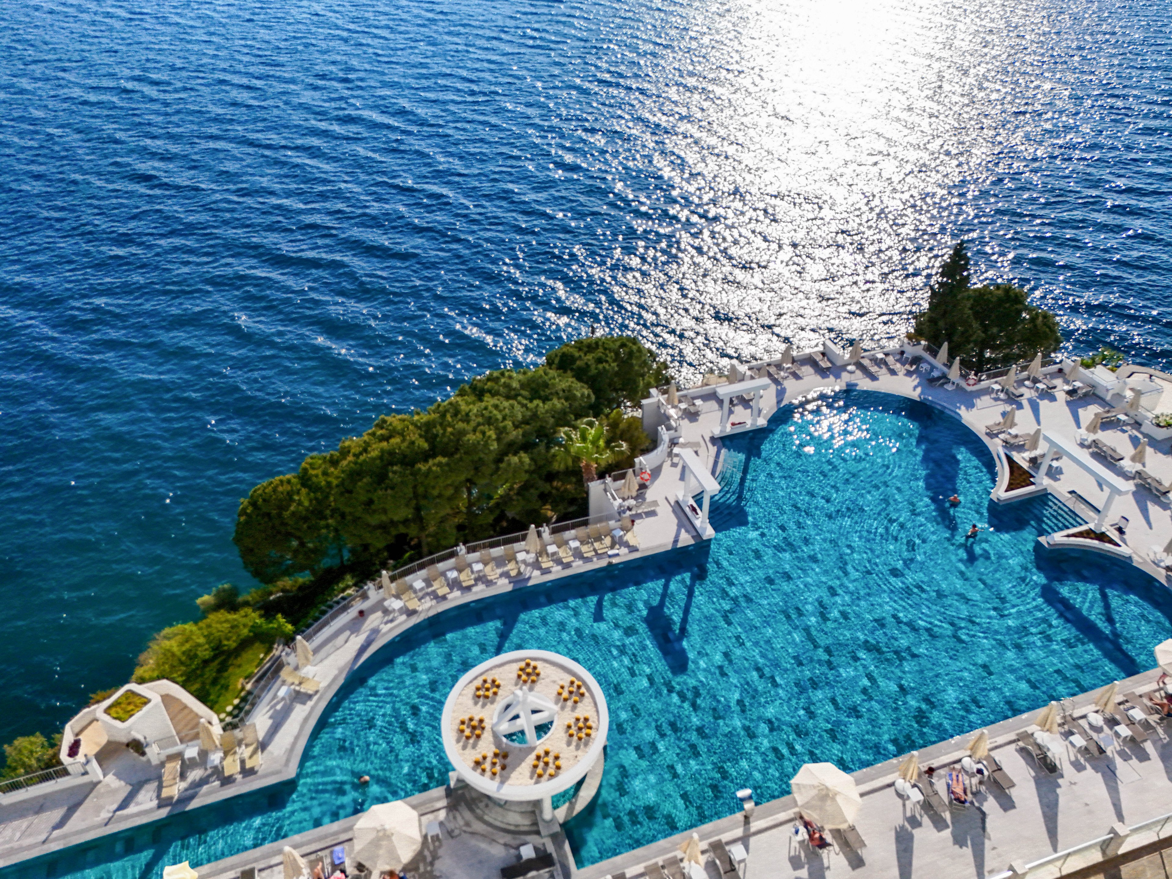A large luxury infinity pool overlooks a calm, expansive sea in this aerial view. White lounge chairs and umbrellas line the poolside terraces, creating a bright, resort-like atmosphere. A circular bar or seating area sits within the pool, adding a focal point to the scene. The sunlight reflects off the blue water, enhancing the tranquil and upscale coastal setting.