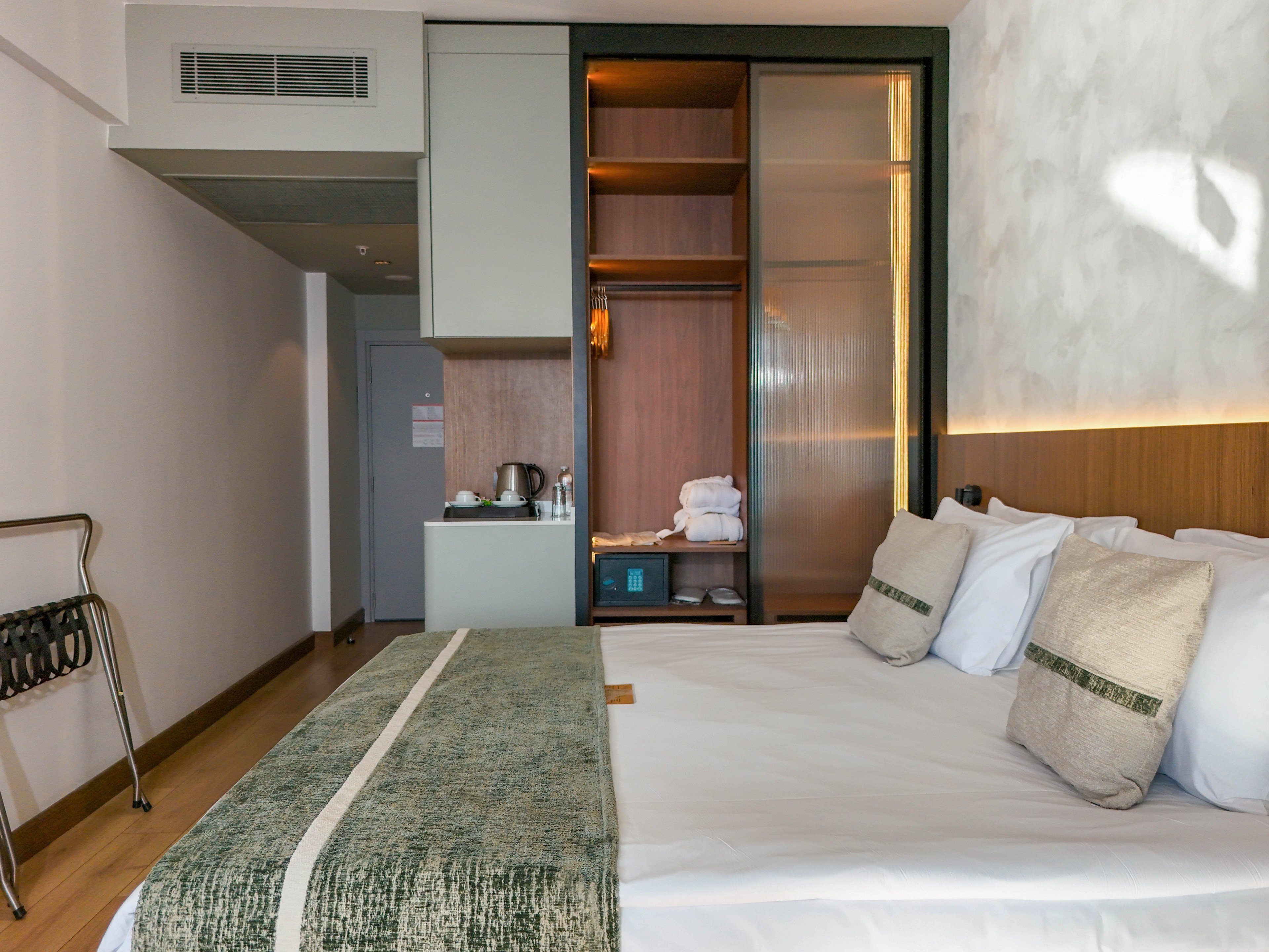 A contemporary hotel room features a neatly made double bed with white linens and textured green accents. The space includes an open wardrobe with shelves, folded towels, and a sliding glass door. A small refreshment area with a kettle and cups sits near the entrance, while a luggage rack stands by the wall. Soft lighting and clean lines create a calm, minimalist atmosphere suitable for travelers.