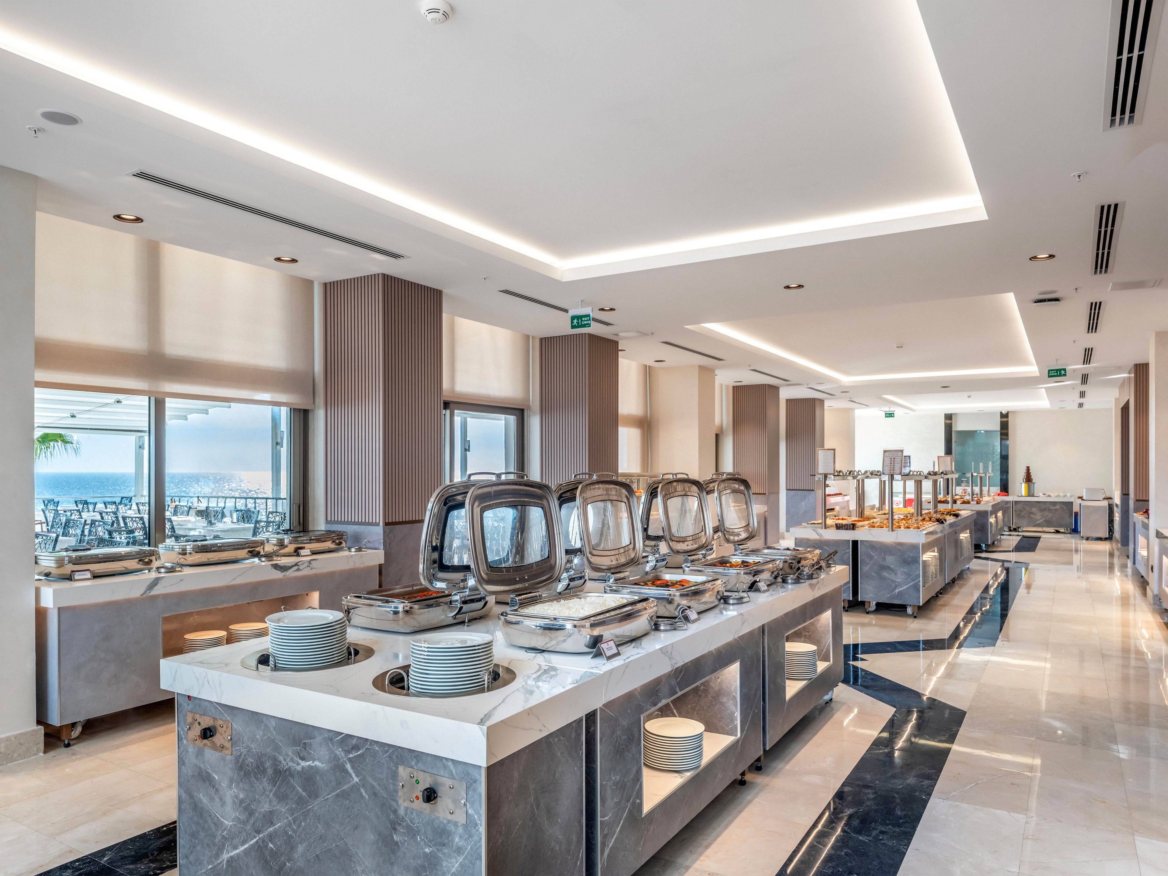 A spacious self-service buffet area is set up in a modern hotel restaurant. Stainless steel chafing dishes and stacks of plates line marble-topped counters. Large windows let in natural light and offer a view of the outdoor seating area. The clean, neutral interior design and bright lighting create a fresh, contemporary dining atmosphere.