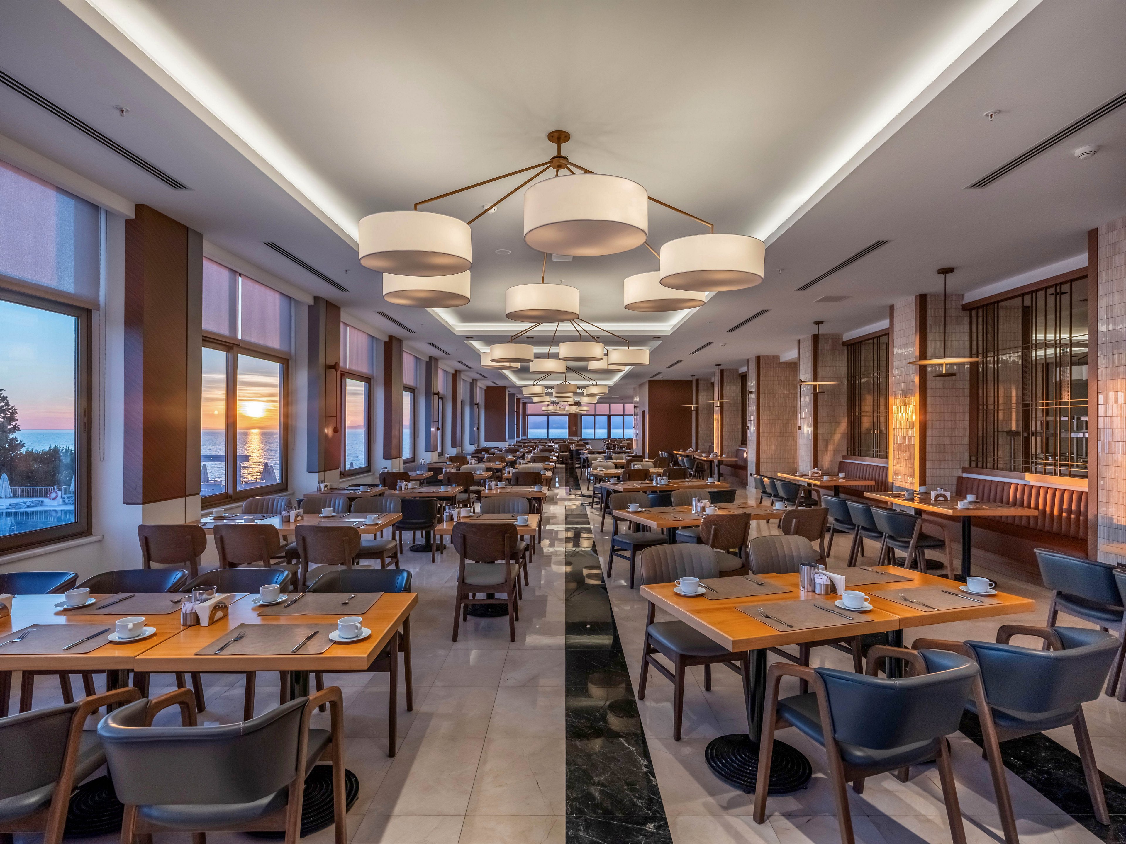 A spacious contemporary restaurant dining room is shown neatly arranged with wooden tables and upholstered chairs. The room is empty of people, emphasizing the orderly place settings with cups, saucers, and napkins. Large windows along one side reveal a colorful sunset sky and reflections on the glass. Warm ceiling lights and soft interior lighting create an inviting, upscale atmosphere.