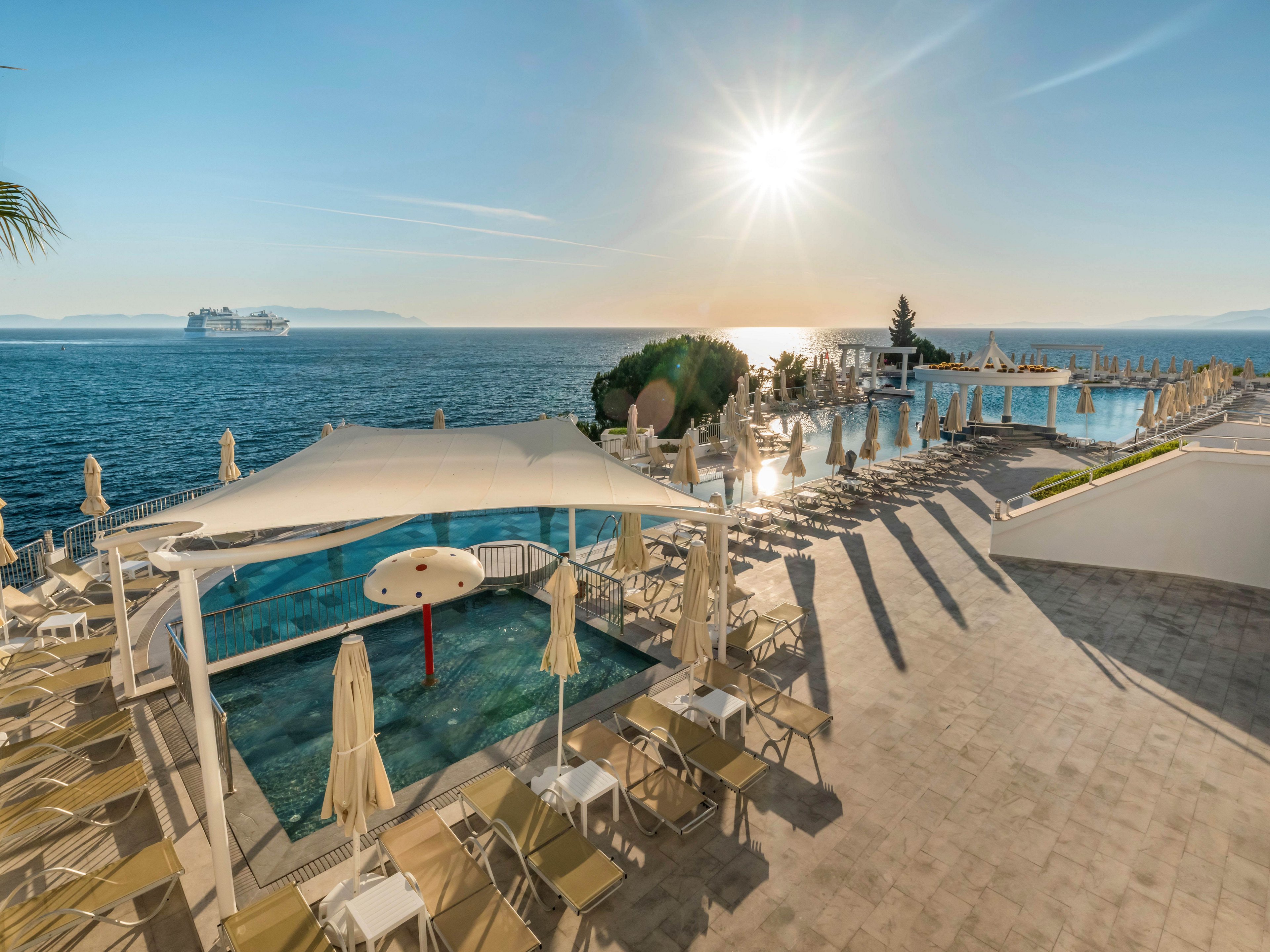A coastal resort features multiple swimming pools overlooking a calm sea at sunset or sunrise. Lounge chairs and umbrellas are neatly arranged around the pools, suggesting a relaxing vacation atmosphere. A shaded kids’ pool area with a mushroom-shaped fountain stands in the foreground. In the distance, a large cruise ship sails across the horizon under a bright, low sun.
