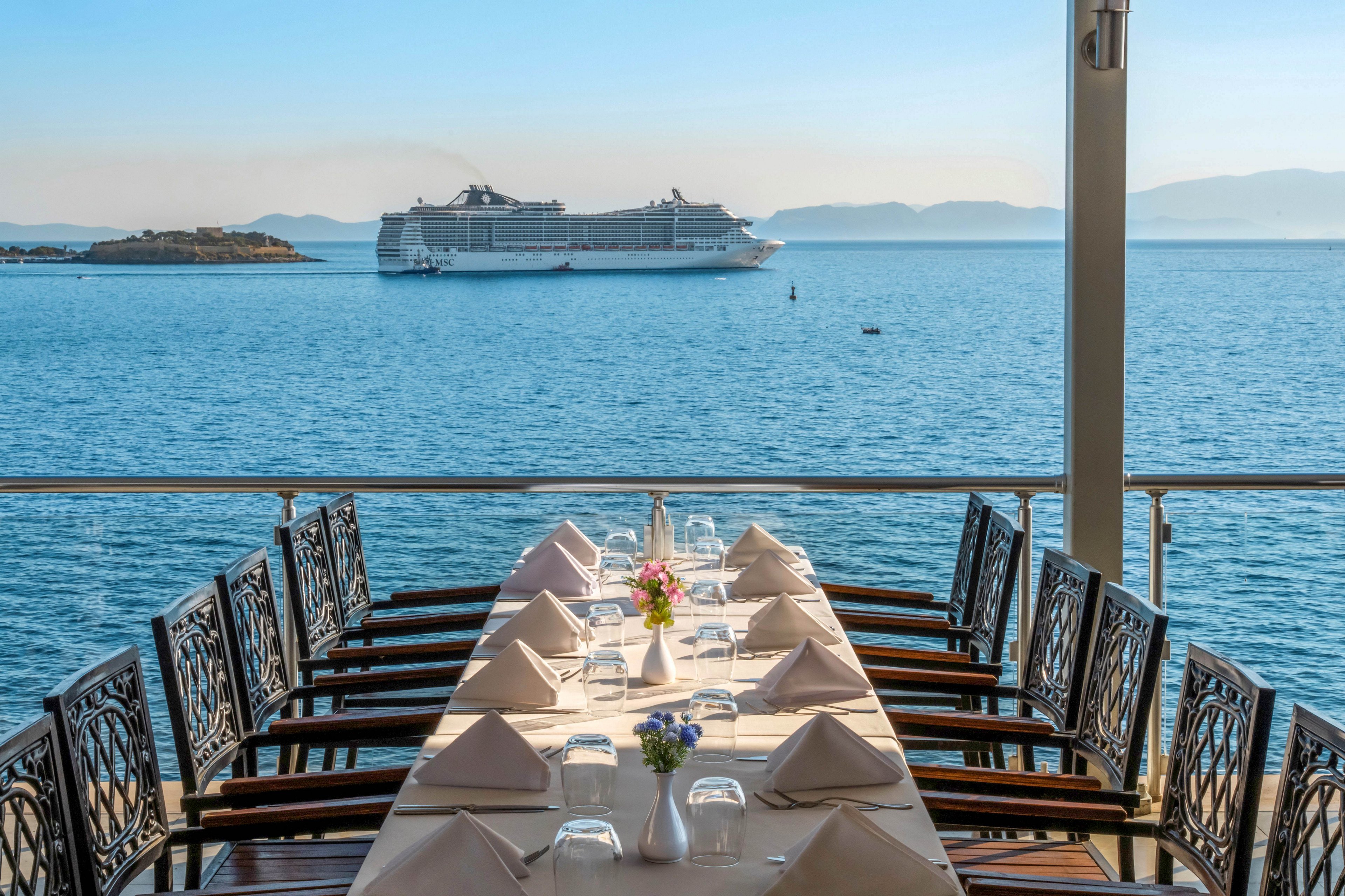 A long outdoor dining table is elegantly set on a seaside restaurant terrace. Folded napkins, glassware, and small flower arrangements decorate the table, ready for guests. Calm blue water stretches into the distance where a large white cruise ship sails past a small island. Soft daylight and clear skies create a relaxed, vacation atmosphere with distant mountains on the horizon.