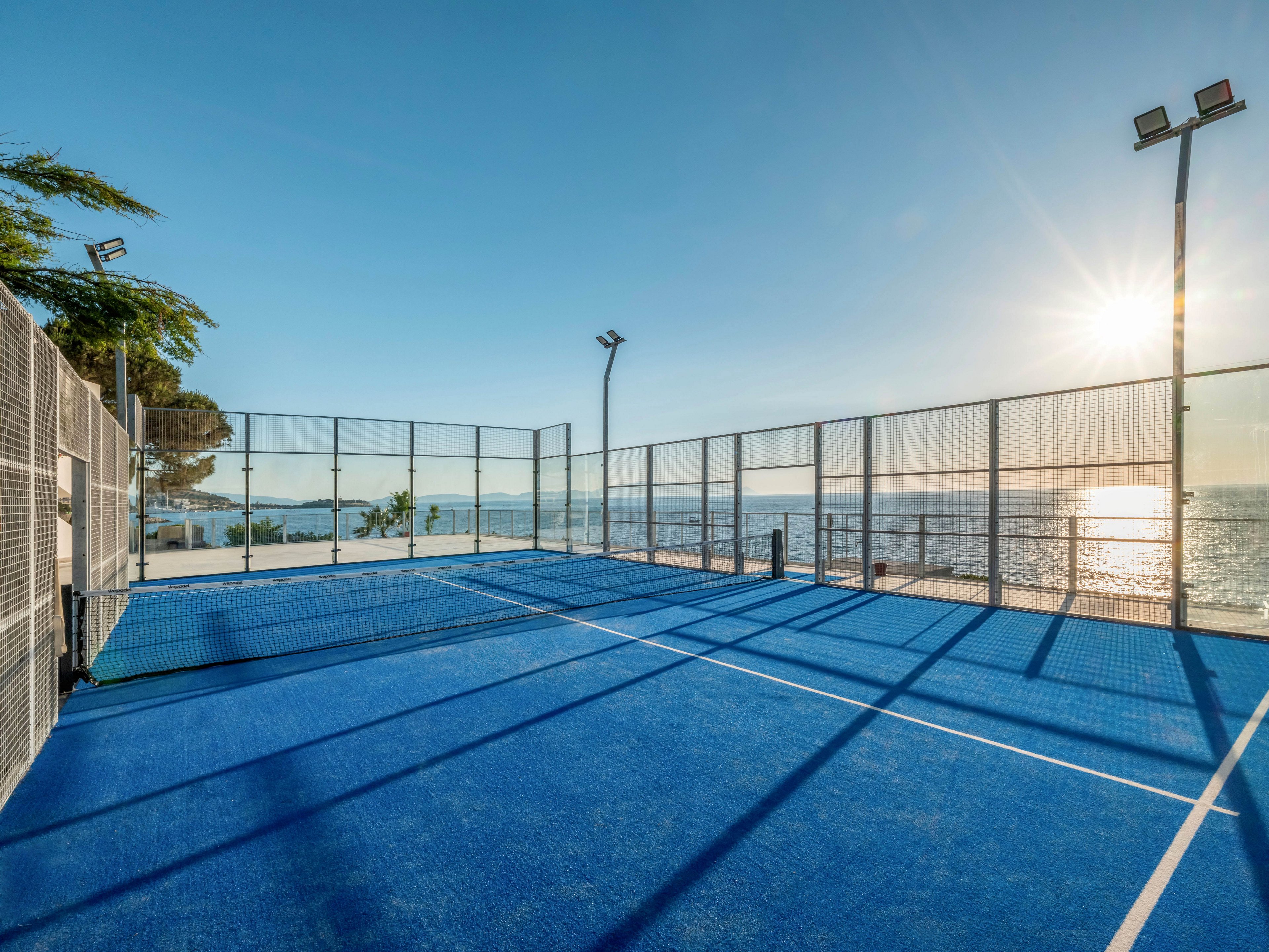 A bright blue padel court stands empty beside the sea under a clear sky. Metal fencing surrounds the court, with tall floodlights positioned at the corners. The low sun casts long shadows across the playing surface and reflects off the calm water. The scene feels peaceful and inviting, suggesting leisure and outdoor sport by the coast.
