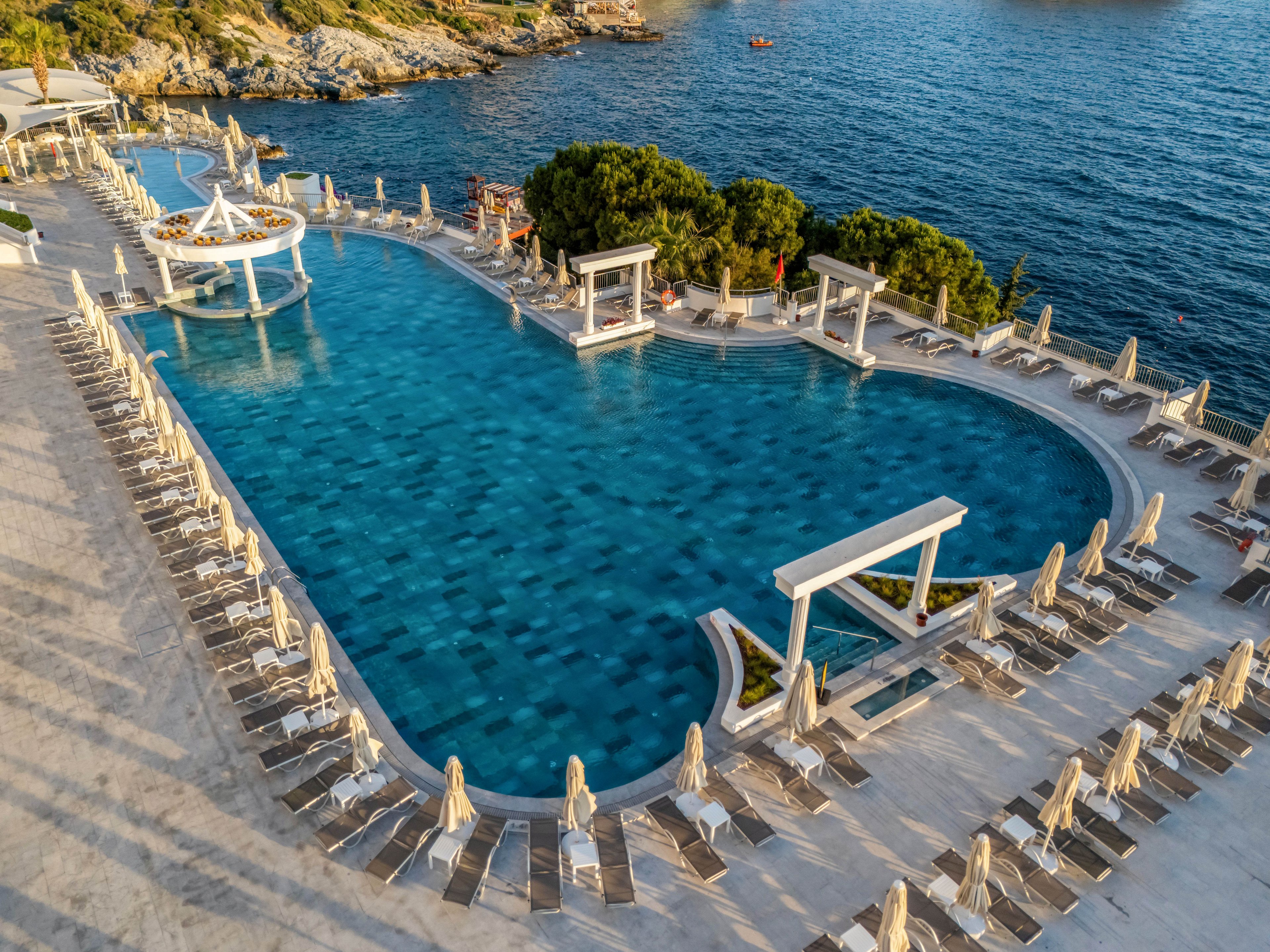A large, elegant infinity pool overlooks a calm blue sea at a coastal resort. Numerous sun loungers with closed umbrellas line the stone pool deck in neat rows. White architectural structures and a circular pool feature add a sophisticated, modern touch. The surrounding rocky shoreline and greenery frame the tranquil waterfront setting under clear daylight.