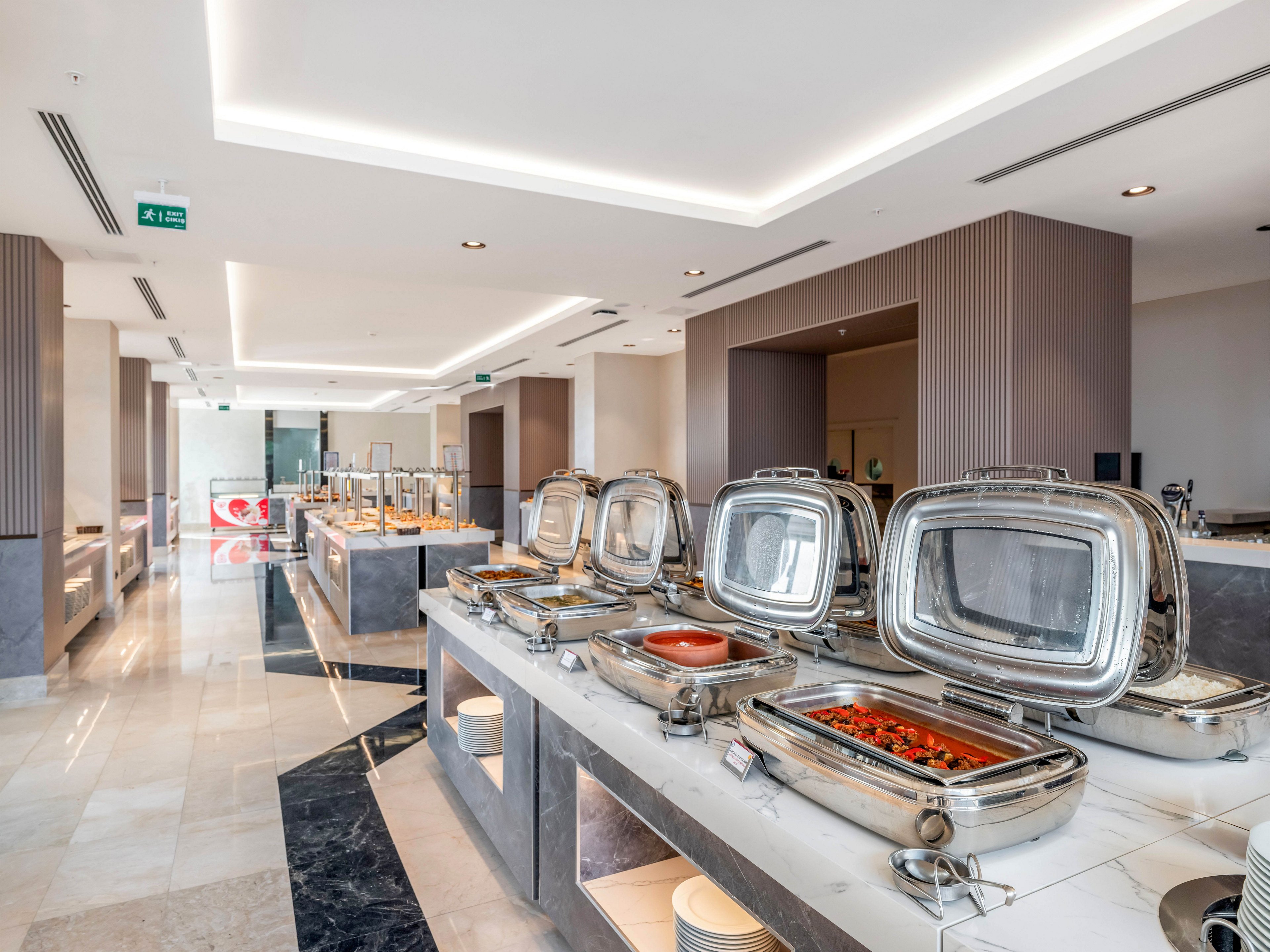 A modern self-service buffet area features a row of stainless steel chafing dishes on a long counter. The space appears bright and spacious with polished floors, recessed ceiling lights, and contemporary architectural details. Some of the buffet trays contain hot food, while others are closed and ready for service. No guests are present, emphasizing the clean, organized presentation of the dining area.