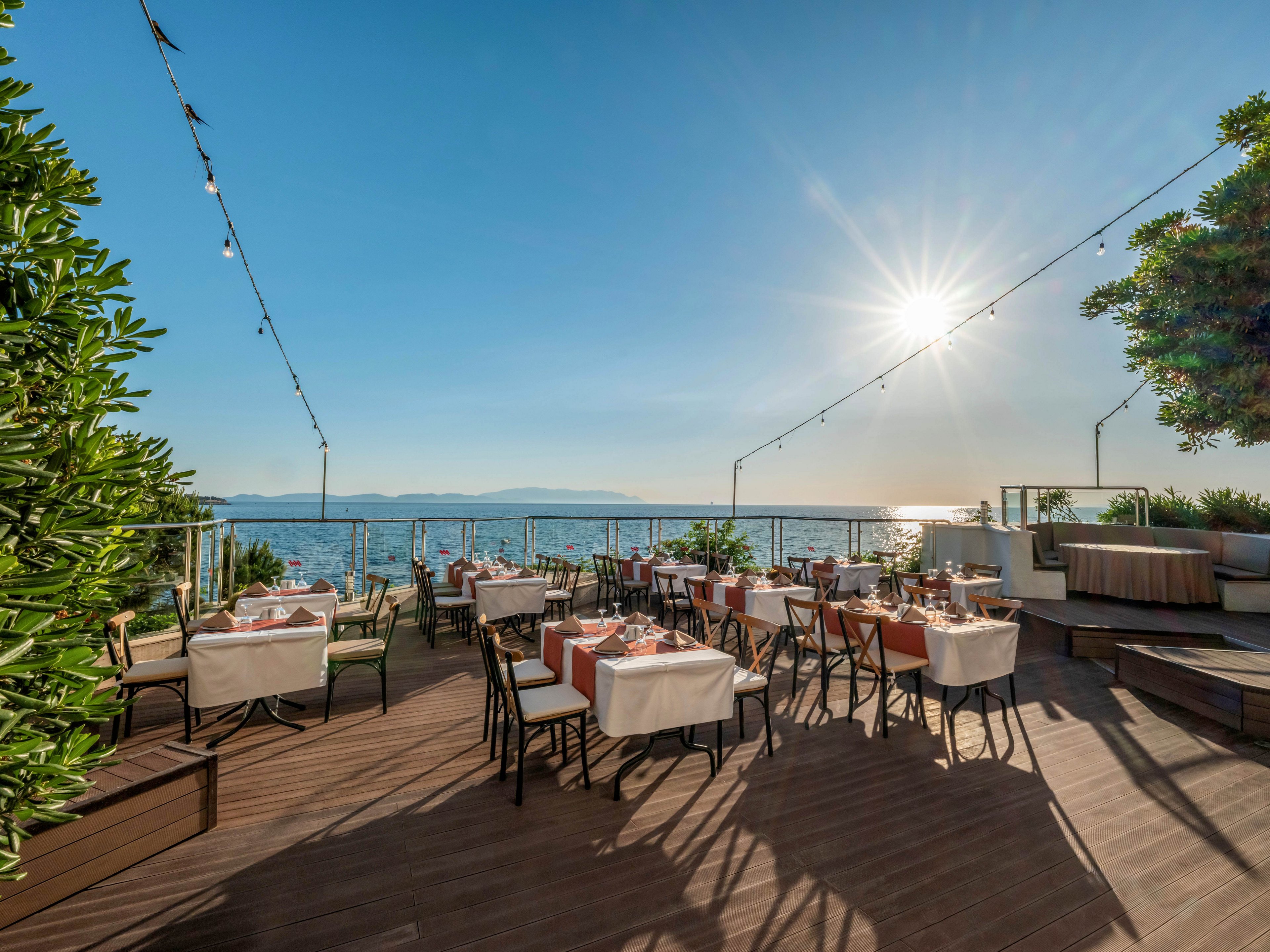 An elegant open-air restaurant terrace overlooks a calm sea under a clear blue sky. Tables are neatly set with white tablecloths, napkins, and glassware, ready for guests. String lights hang above the wooden deck, adding a festive touch to the coastal setting. The low sun creates warm reflections on the water and highlights the surrounding greenery and modern railings.