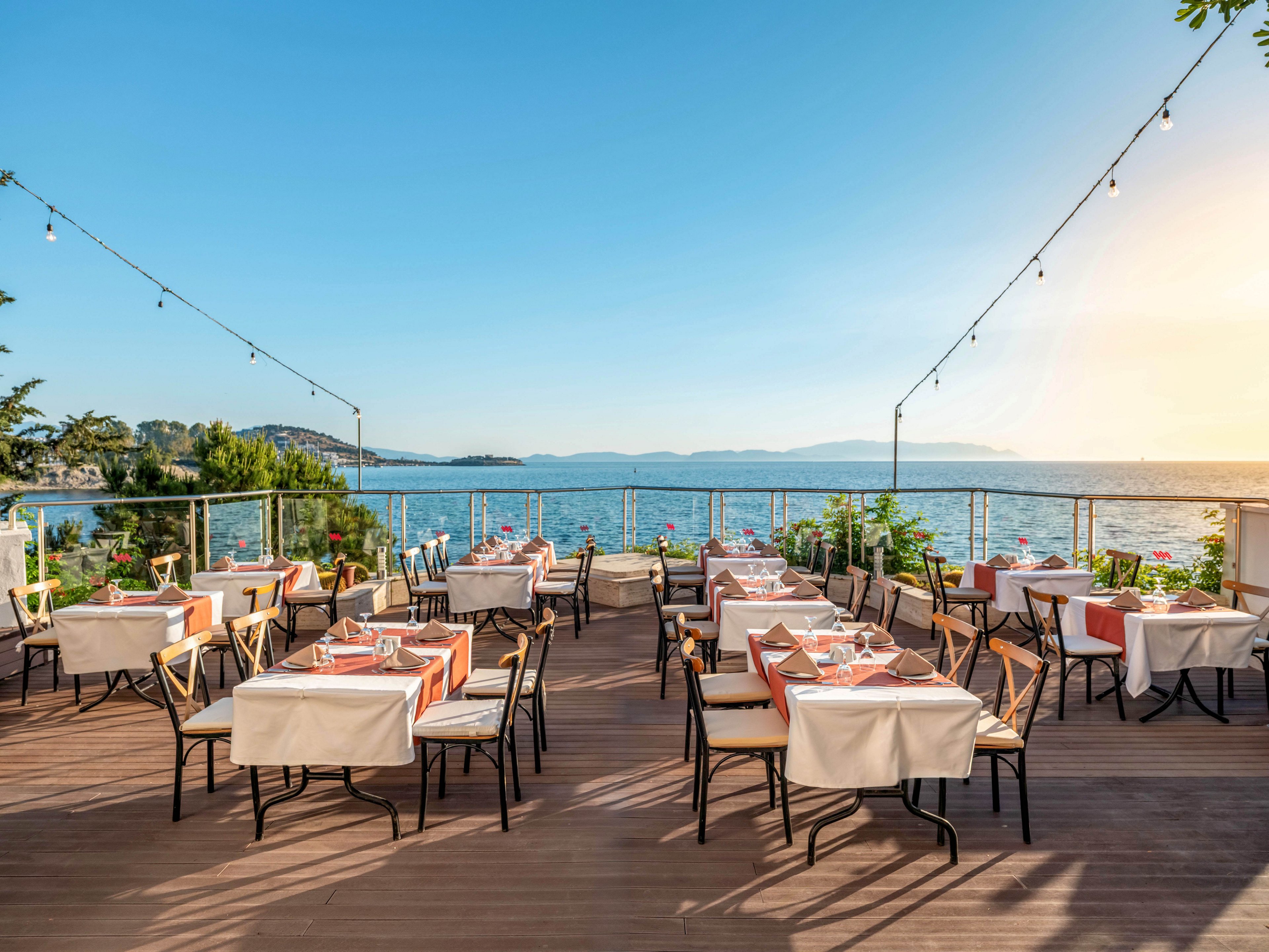 An elegant open-air restaurant terrace overlooks a calm sea under a clear blue sky. Tables are neatly set with white tablecloths, napkins, and glassware, ready for guests. String lights are suspended above the wooden deck, adding a warm and inviting ambiance. Lush greenery and a glass railing frame the view of distant mountains and the water.