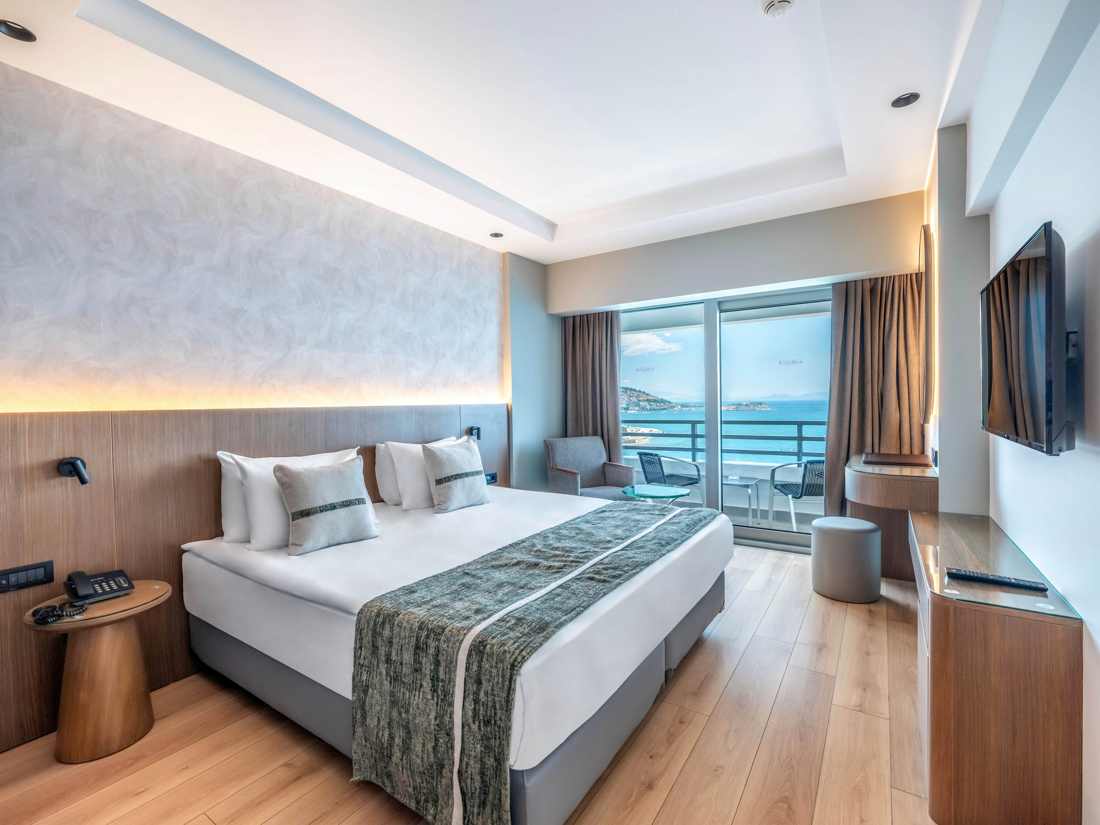 Contemporary hotel room featuring a large neatly made bed with decorative pillows and runner. The space includes wooden furniture, a wall-mounted television, and a small seating area. Floor-to-ceiling glass doors open to a balcony with chairs overlooking the sea and distant coastline. Soft neutral tones and natural light create a calm, inviting atmosphere suitable for relaxation or vacation stays.