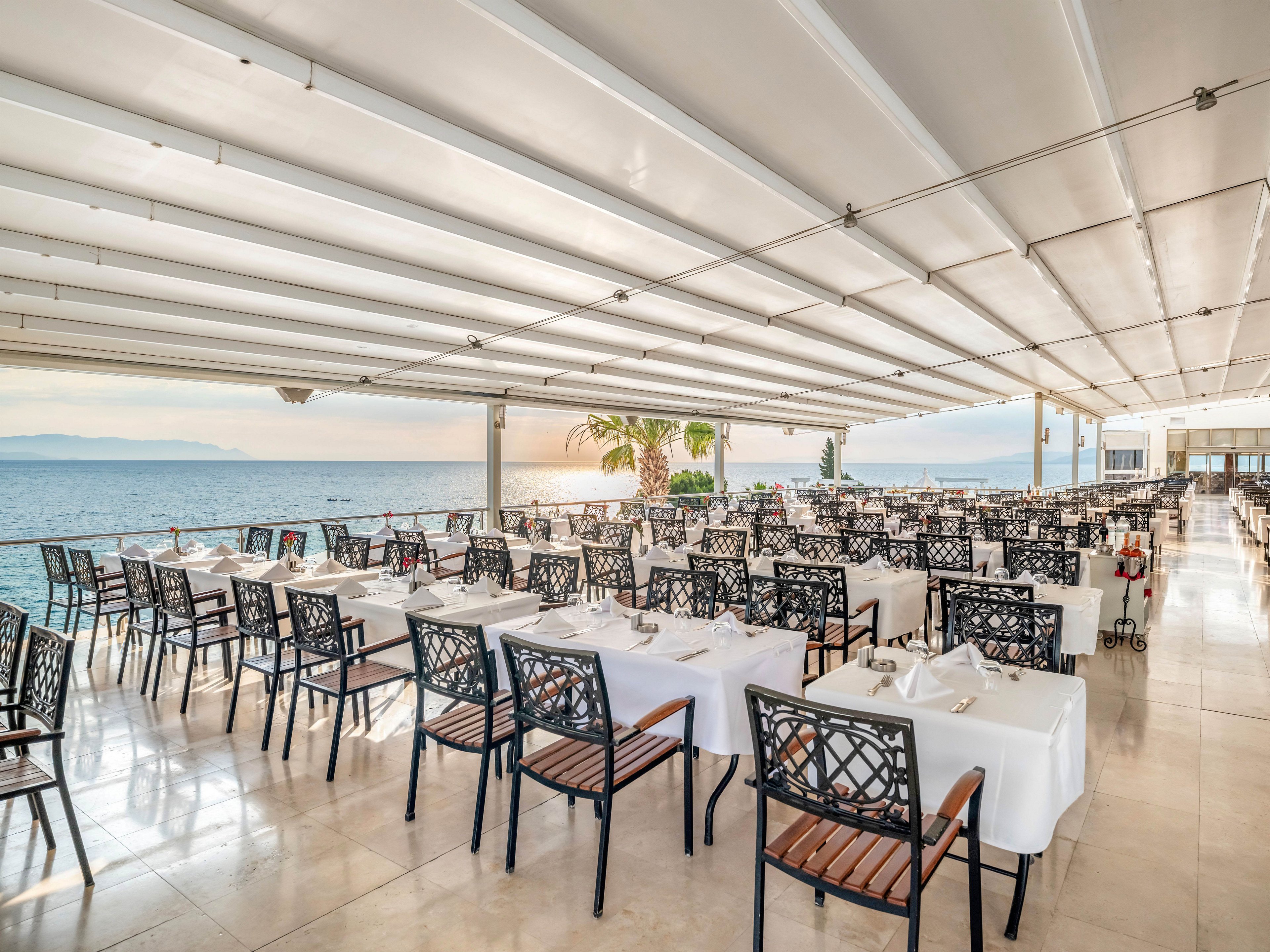 An elegant open-air restaurant is set with rows of white tablecloth-covered tables and black metal chairs. The dining area sits under a modern covered terrace overlooking a calm sea. Soft sunset light reflects on the polished floor, creating a warm and inviting atmosphere. The scene is empty of people, emphasizing the spacious layout and serene coastal setting.