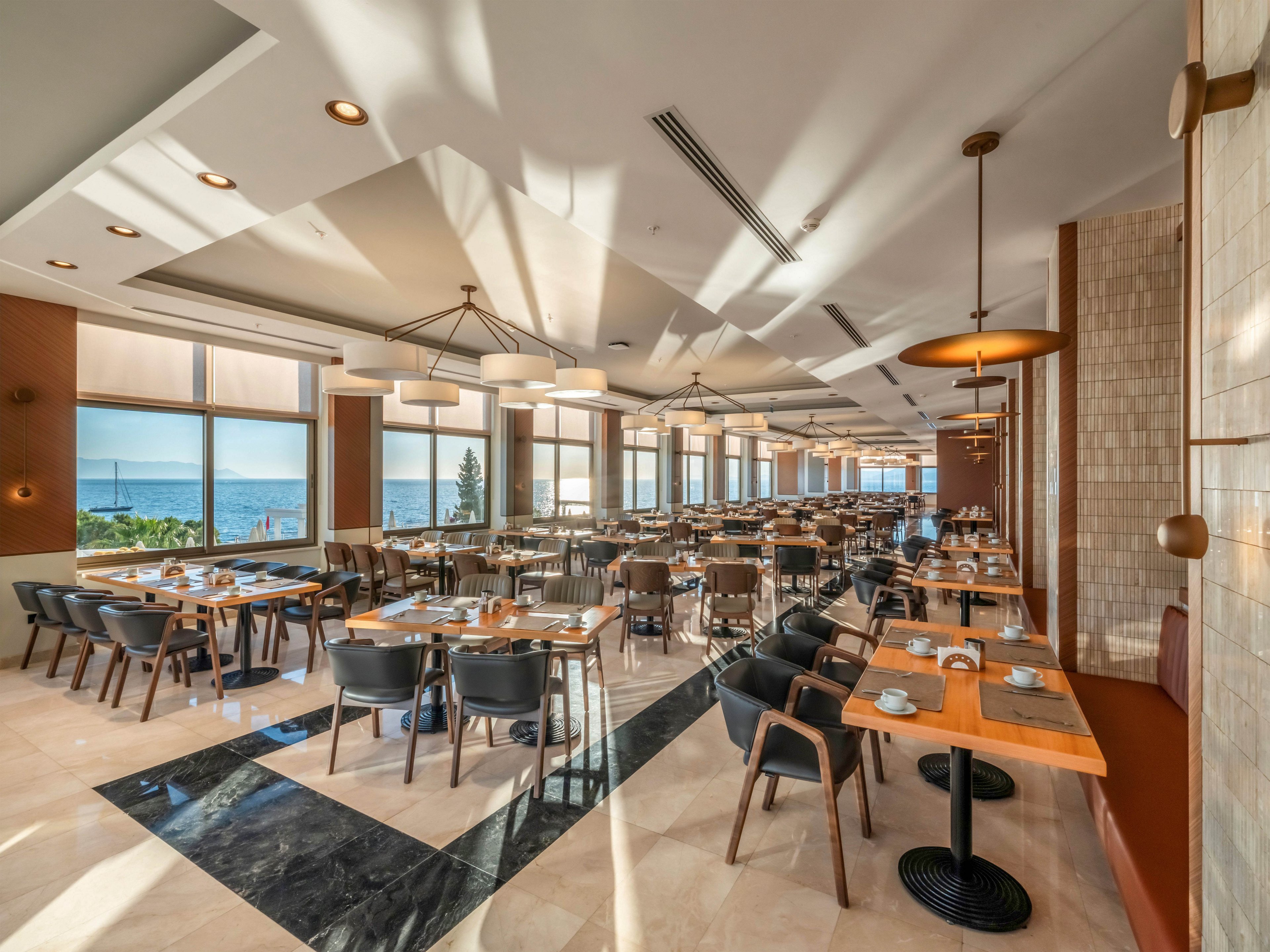 Spacious contemporary restaurant interior with neatly arranged wooden tables and chairs. Large floor-to-ceiling windows let in abundant natural light and reveal a scenic sea view and greenery outside. Warm neutral tones, geometric floor patterns, and stylish pendant lighting create an inviting dining atmosphere. Table settings with plates, cups, and cutlery suggest readiness for guests in a calm, empty space.
