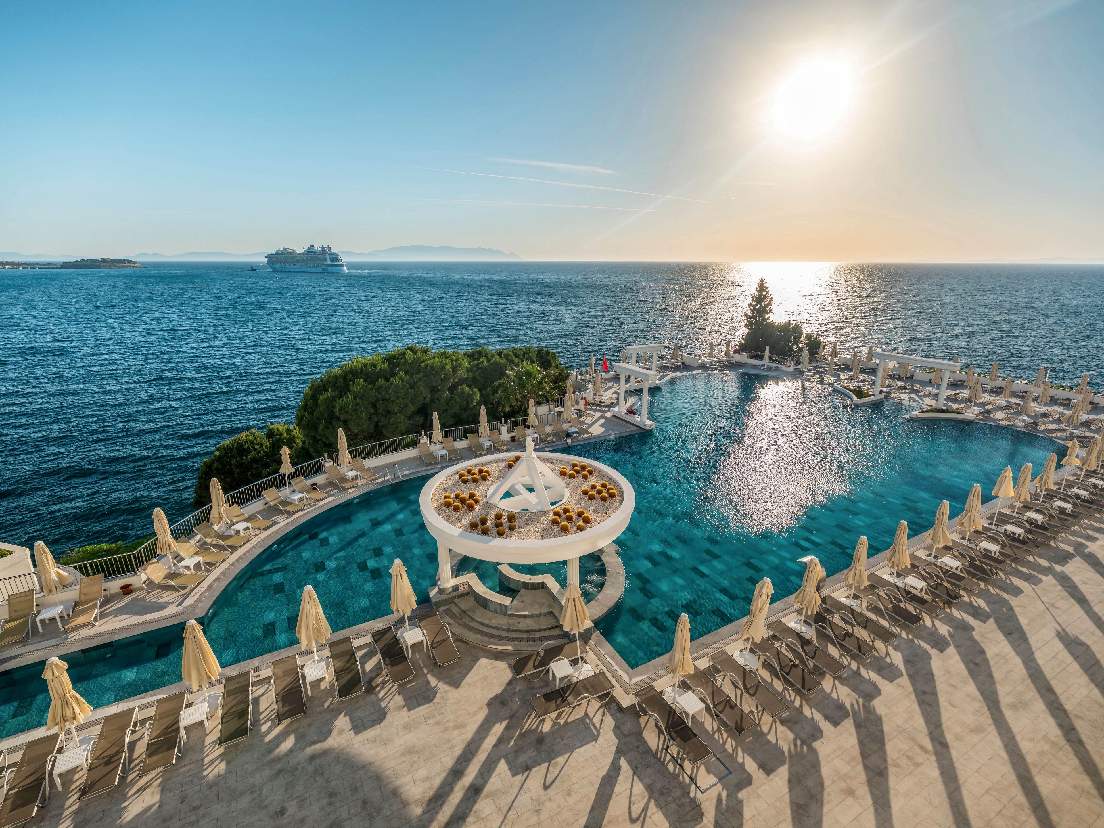 A large, curved swimming pool overlooks a calm, expansive sea under a bright setting sun. Numerous sun loungers and umbrellas surround the pool, creating a relaxing resort atmosphere. A circular pool bar or seating area sits at the center of the water, adding a focal point to the scene. In the distance, a cruise ship sails across the horizon, enhancing the sense of vacation and coastal escape.