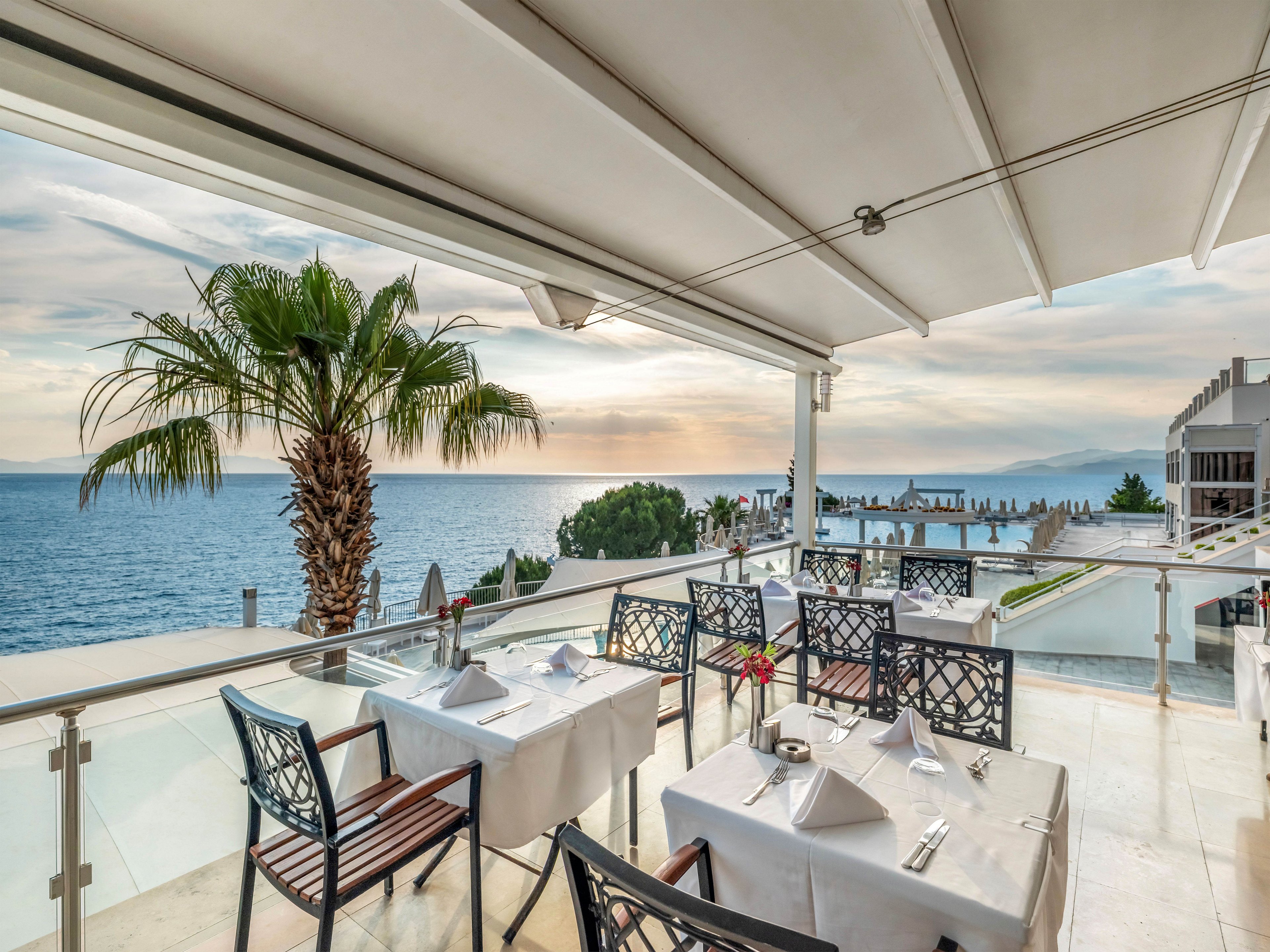 An elegant outdoor restaurant terrace overlooks a calm ocean at sunset. White tablecloths, neatly arranged place settings, and dark metal chairs create a refined dining atmosphere. A tall palm tree and glass railing frame the expansive water and distant mountains. The soft evening light and pastel sky give the scene a tranquil, romantic mood.