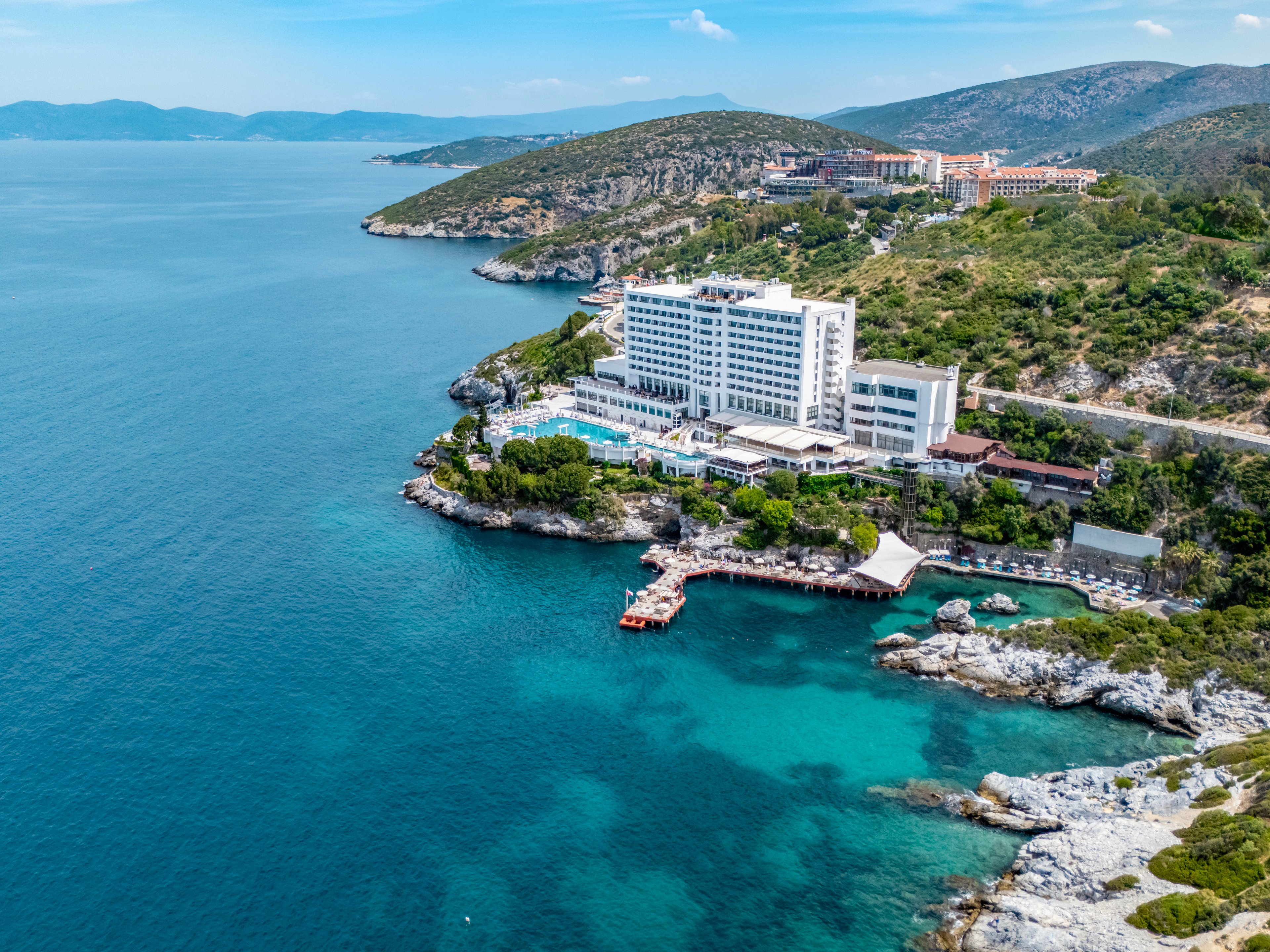 An aerial view shows a large seaside resort hotel built on a rocky coastline. The white multi-story building sits beside a swimming pool complex and terraces surrounded by greenery. Clear turquoise water fills the sheltered bay below, contrasting with the deeper blue of the open sea. Rolling hills and additional coastal developments stretch into the distance under a bright, clear sky.