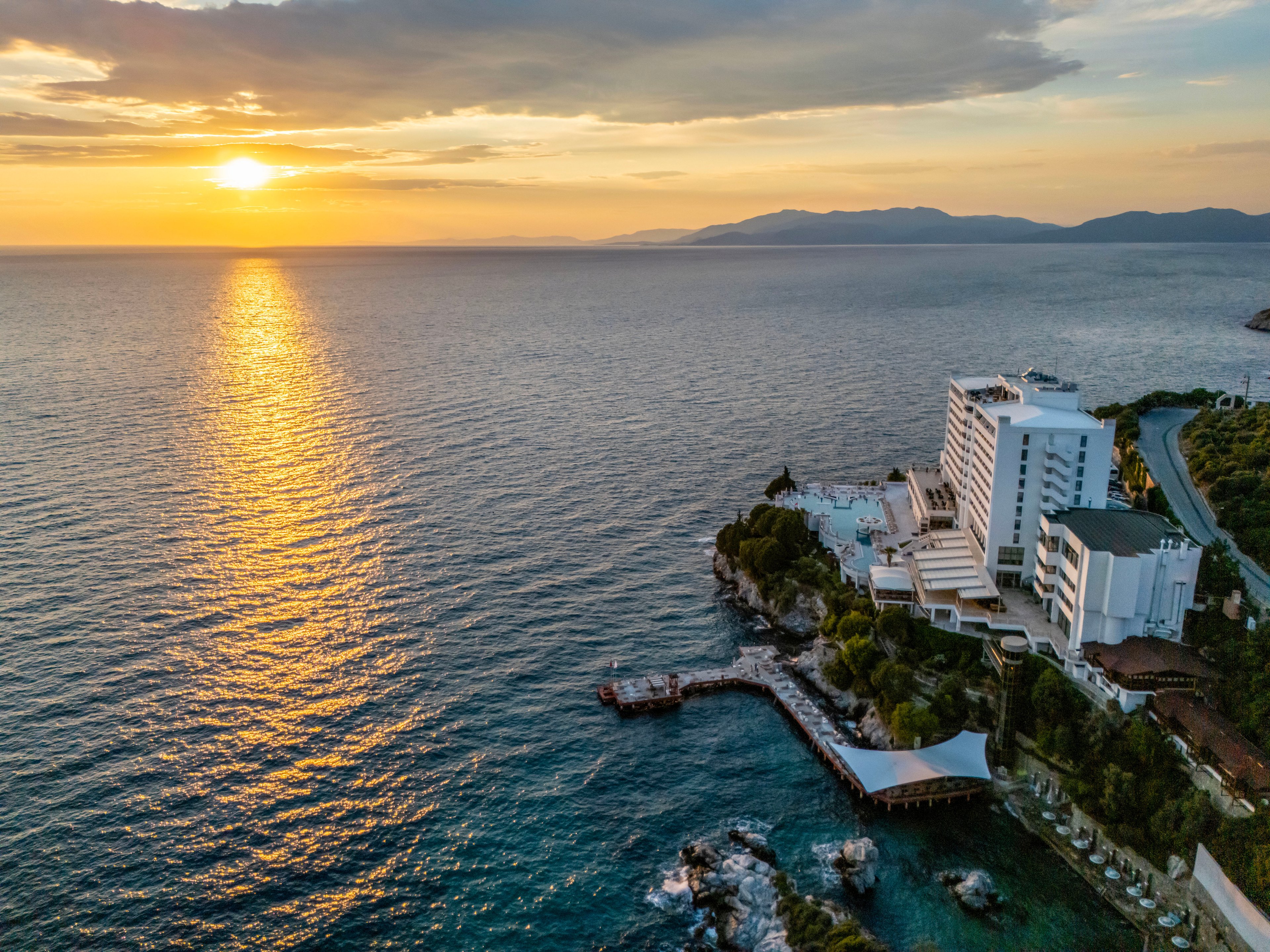 An aerial view captures a modern seaside resort hotel perched on a rocky coastline at sunset. The sun is low on the horizon, casting golden light across the calm sea and creating a shimmering reflection on the water. The hotel complex includes multiple white buildings, terraces, and a pool area surrounded by greenery. A coastal road curves along the shoreline, connecting the resort to the distant mountainous landscape under a partly cloudy sky.