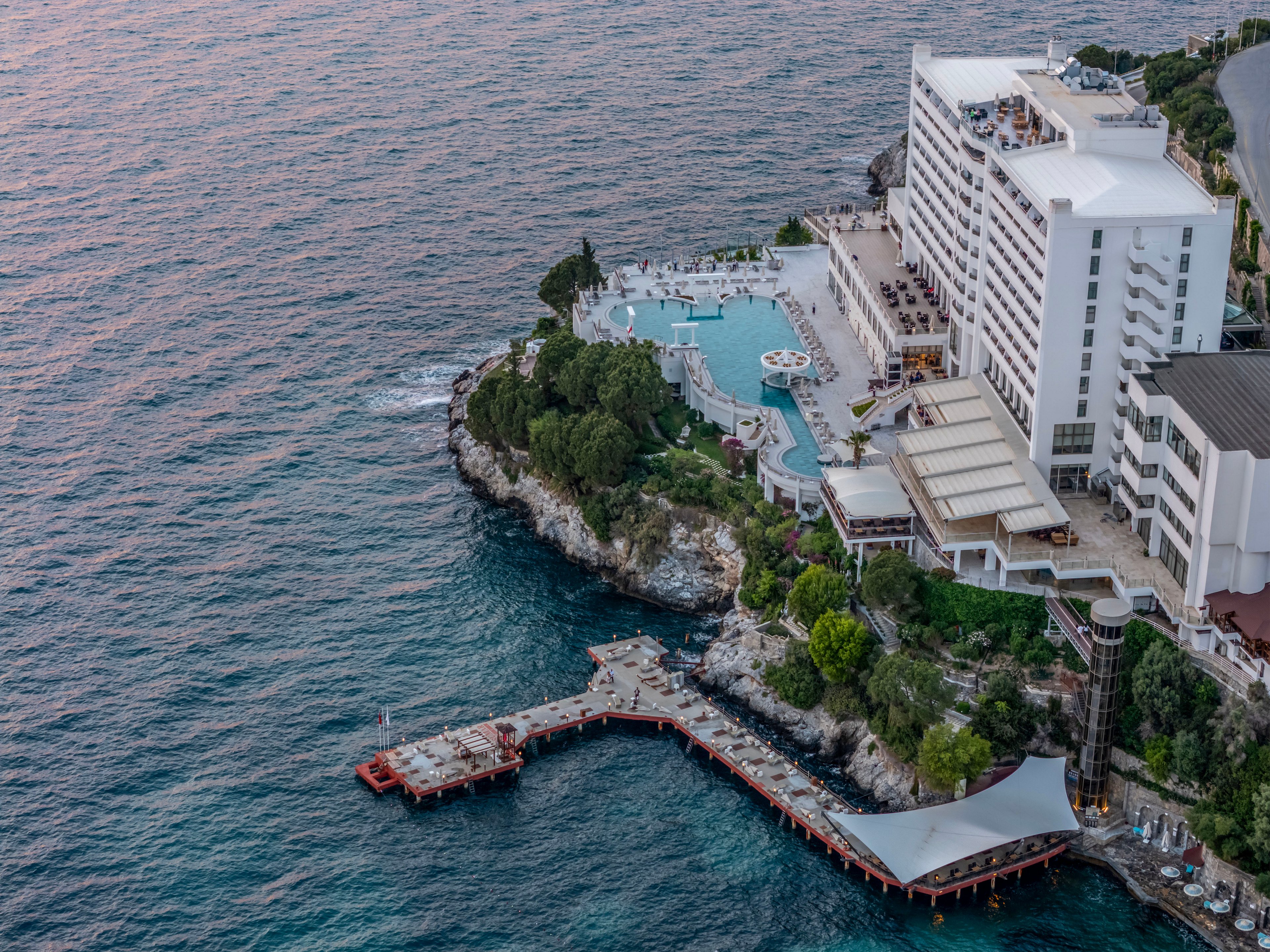 A large waterfront resort sits dramatically on a rocky cliff overlooking the sea. The complex features a spacious outdoor swimming pool area surrounded by sun loungers and terraces. Below, a wooden pier and platforms extend over the water, providing direct access to the ocean. Lush green trees and shrubs soften the rocky shoreline, while calm waves and soft evening light create a tranquil coastal atmosphere.