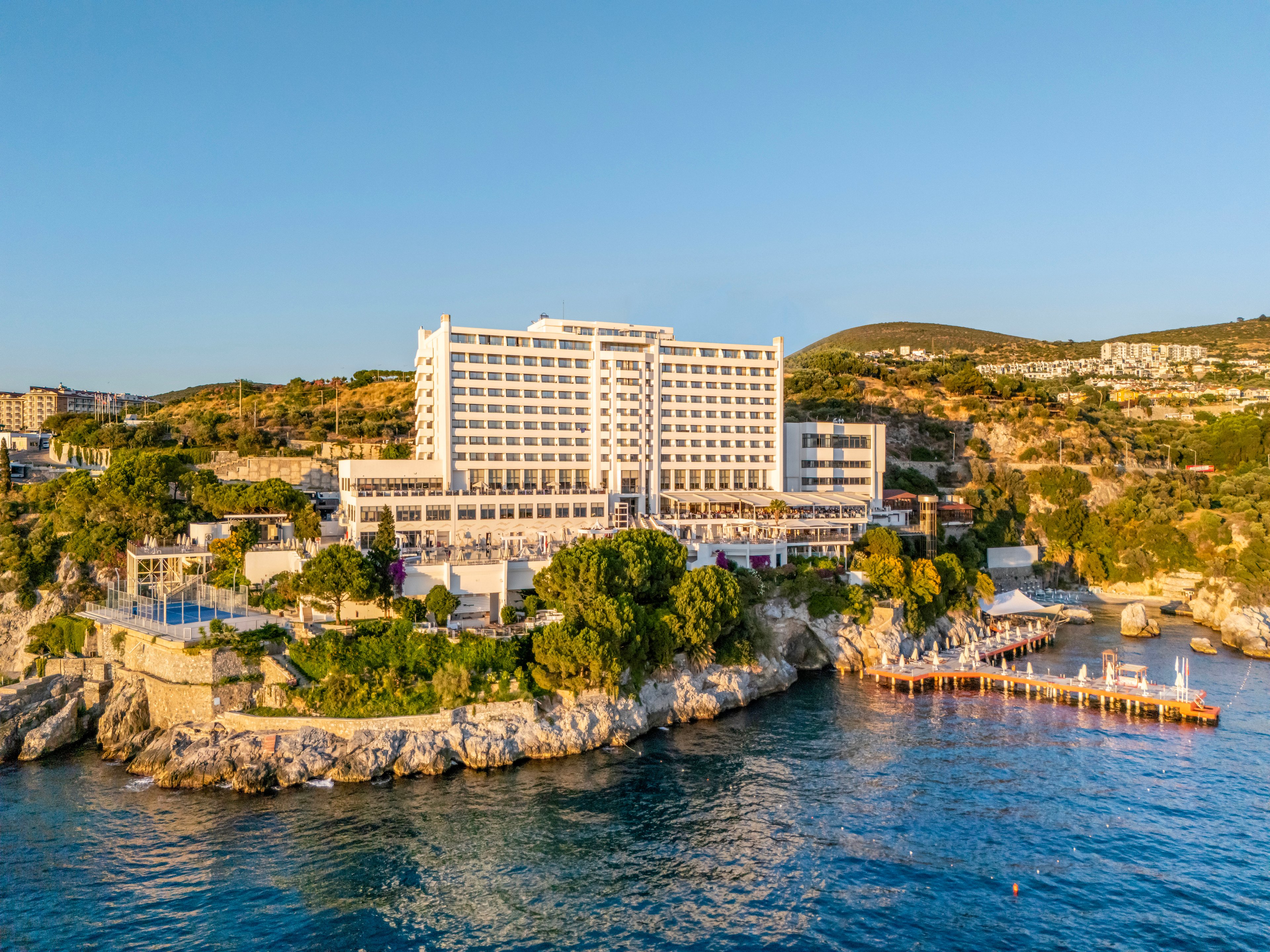 A large multi-story resort hotel stands prominently on a rocky shoreline overlooking calm blue sea waters. The building is surrounded by greenery and terraced areas leading down to the water. Wooden piers and sun decks extend into the sea, suggesting leisure and vacation activities. Hills with scattered buildings rise in the background under a clear golden-hour sky.