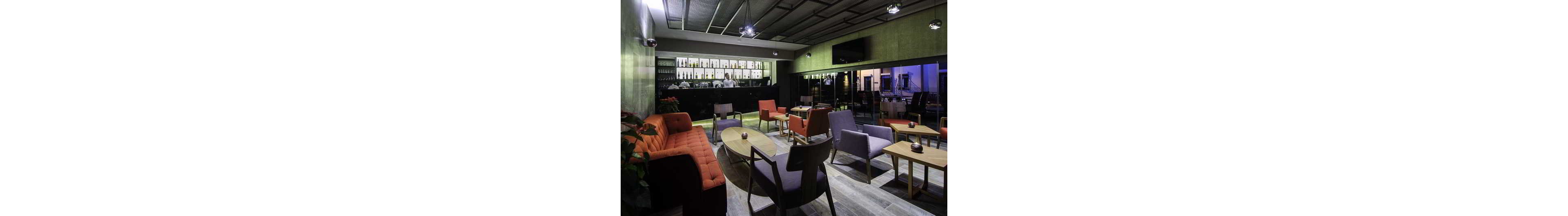 A contemporary lounge bar features a mix of sofas, armchairs, and small tables arranged in an open seating area. The bar counter at the back displays bottles and glassware, with a bartender standing behind it. Large windows on one side reveal an urban night view with blue and purple lighting. The overall atmosphere is relaxed and stylish, with muted tones accented by orange and red chairs.