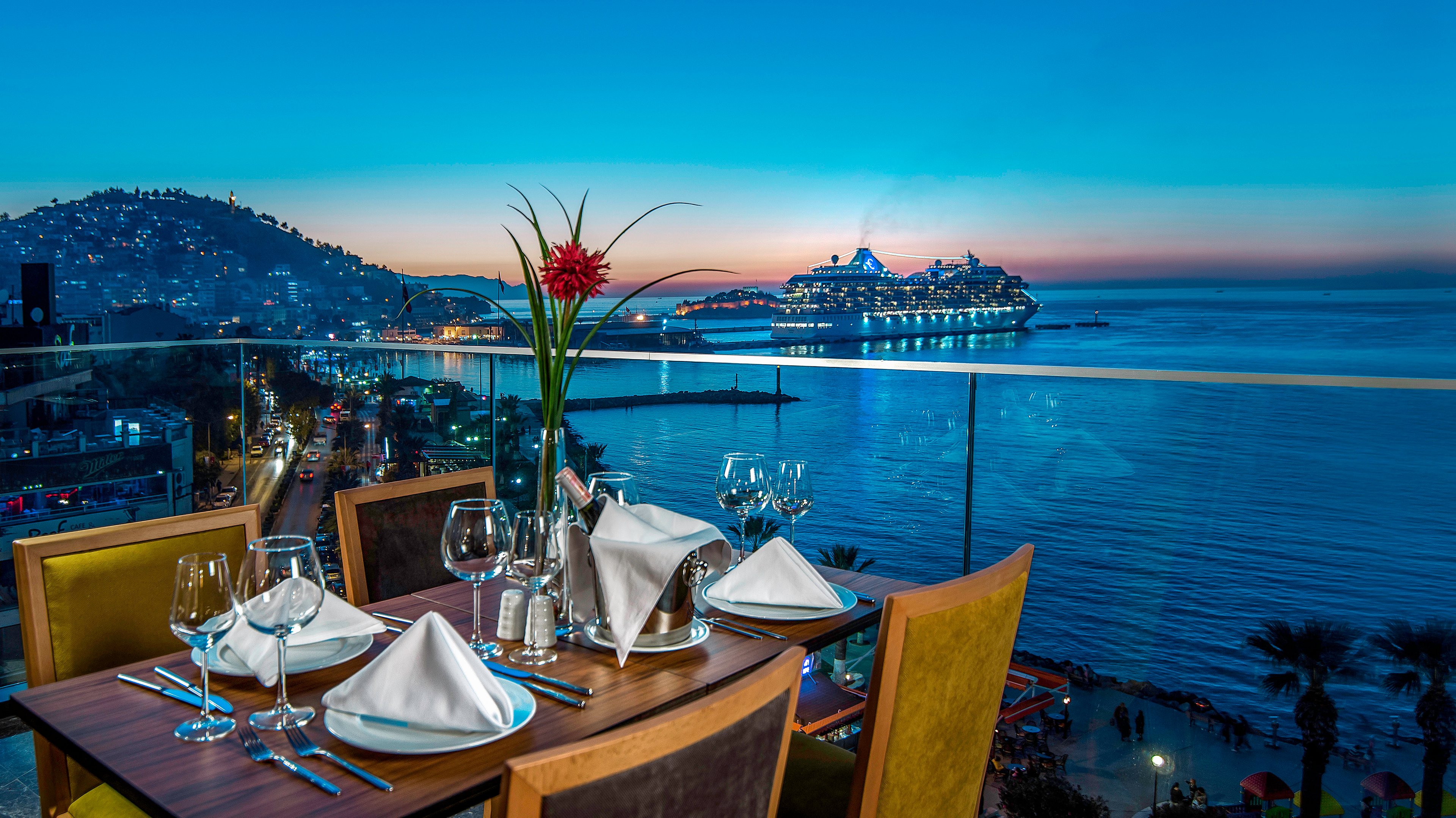 An elegant restaurant table is set for dinner on a balcony overlooking a calm seaside bay. White napkins, wine glasses, and a single red flower centerpiece create a refined dining atmosphere. Below, a coastal city with hillside lights stretches along the shoreline as evening falls. In the distance, a brightly lit cruise ship is anchored on the water under a vivid blue twilight sky.