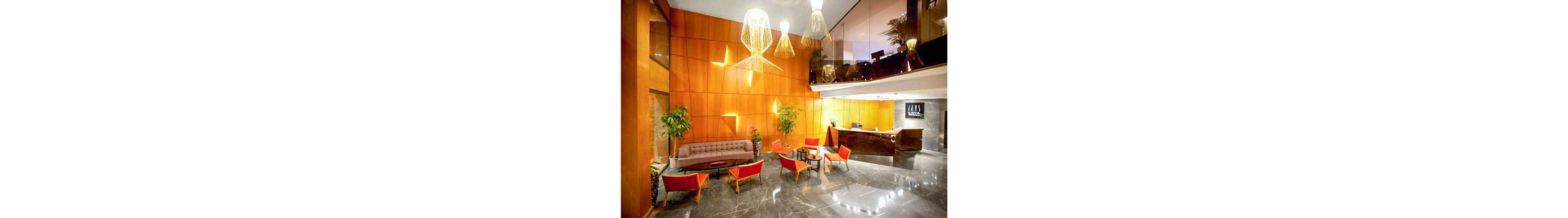 A contemporary hotel lobby features a sleek reception desk and a stylish seating area. Warm wooden wall panels contrast with glossy dark marble flooring. Bright orange chairs and a neutral sofa surround small round tables, creating an inviting space for guests. Large pendant lights hang from the ceiling, and potted plants add greenery to the modern interior.