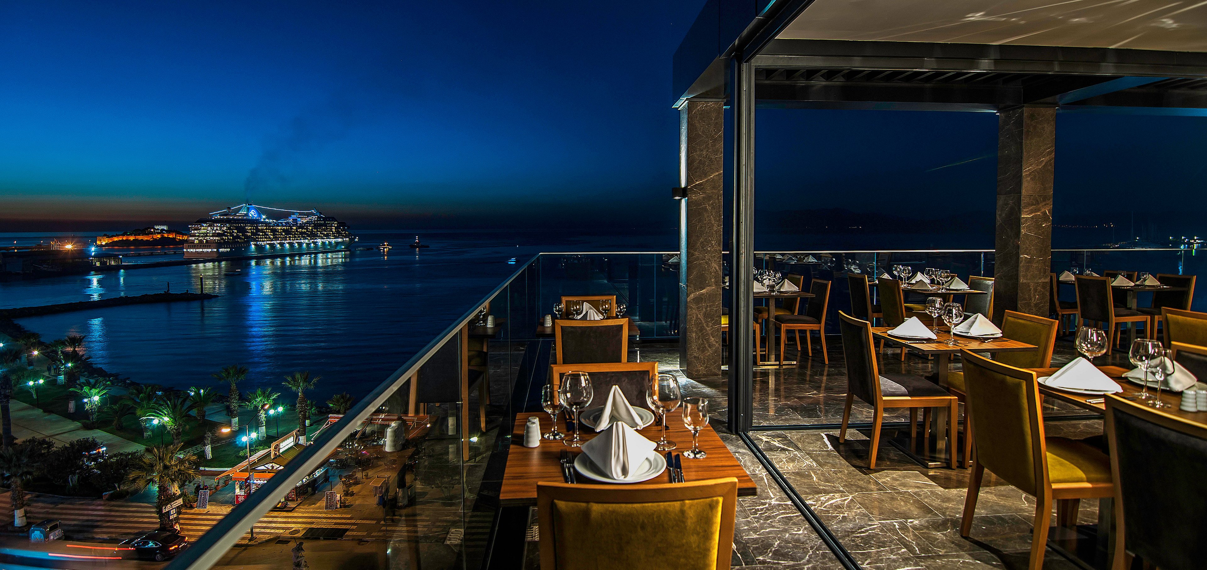 A stylish rooftop restaurant terrace overlooks a calm harbor at night. Set tables with folded white napkins and glassware line the edge of the balcony, ready for evening diners. In the distance, a brightly lit cruise ship glows against the deep blue sky and water. City lights and palm trees illuminate the shoreline, creating a sophisticated and romantic coastal ambiance.