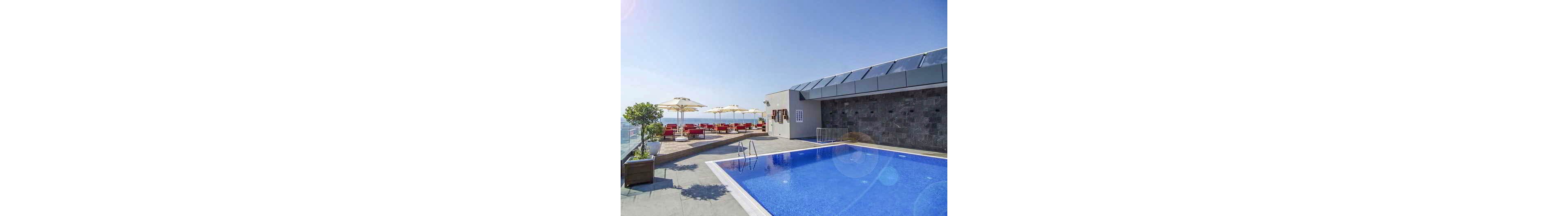 A modern rooftop swimming pool overlooks a distant ocean horizon under a clear blue sky. The pool area features clean lines, bright blue water, and a stone wall along one side. In the background, a terrace with red seating and white umbrellas offers a relaxing lounge space. Potted plants and sunlit reflections contribute to a calm, resort-like atmosphere.