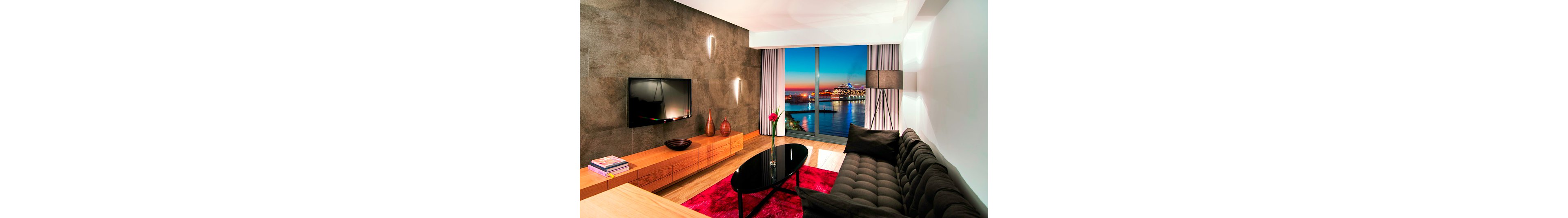 A contemporary living room features a dark tufted sofa, sleek black coffee table, and low wooden media console. Large floor-to-ceiling windows reveal a colorful waterfront cityscape at dusk. Warm accent lighting, decorative vases, and a bright red rug add contrast to the neutral walls and wood flooring. A flat-screen television is mounted on the textured wall, completing the modern interior design.