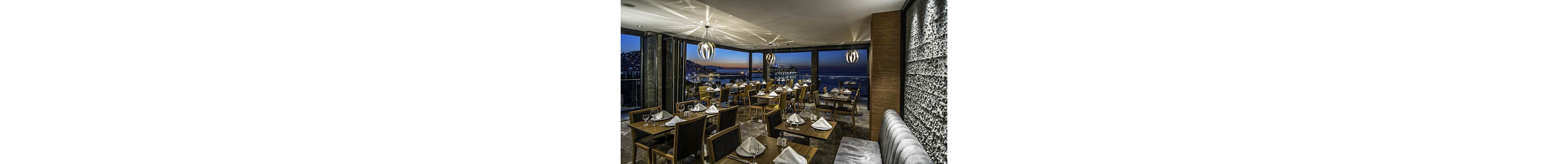 A contemporary restaurant dining room is set with neatly arranged wooden tables and cushioned chairs, each place setting prepared with folded white napkins and glassware. Large floor-to-ceiling windows reveal a coastal cityscape and calm sea at twilight, with lights twinkling along the shoreline. Stylish pendant lights hang from the ceiling, casting a warm glow over the polished floor and textured wall. The atmosphere feels elegant yet relaxed, ideal for evening dining with a scenic ocean view.