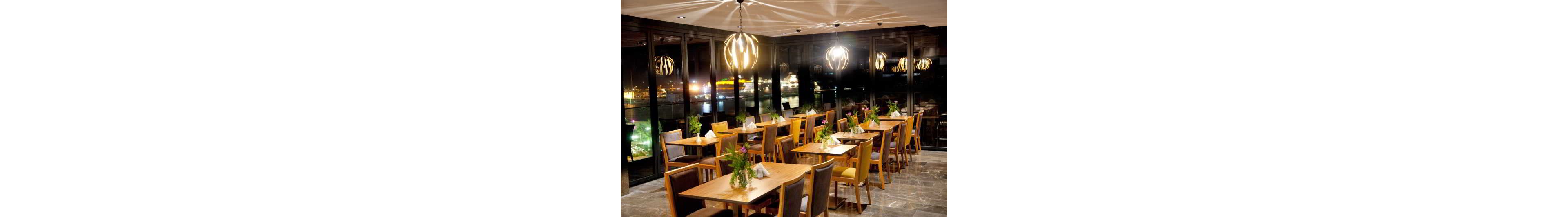 A contemporary restaurant dining area features neatly arranged wooden tables and chairs, each decorated with small floral centerpieces and folded napkins. Large floor-to-ceiling windows reveal a brightly lit harbor or cityscape at night, adding a scenic backdrop to the interior. Warm pendant lights hang from the ceiling, casting a cozy glow over the polished floor and furniture. The overall atmosphere feels inviting, elegant, and ideal for evening dining with a view.