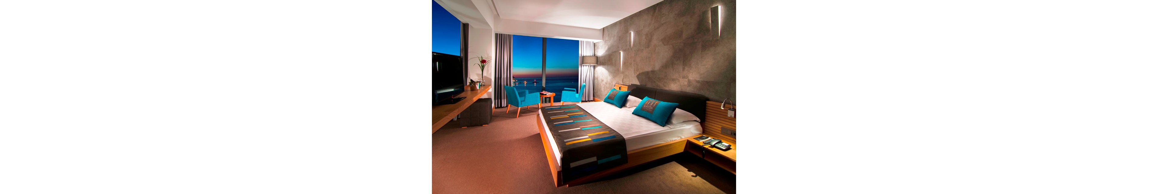 A contemporary hotel bedroom features a large bed with colorful geometric bedding and teal accent pillows. Floor-to-ceiling windows reveal a vivid ocean horizon at sunset, casting a warm glow across the room. A seating area with blue armchairs and a small table is positioned by the window, while a flat-screen TV and sleek wooden furniture complete the space. Soft wall lighting and neutral tones create a calm, upscale atmosphere.