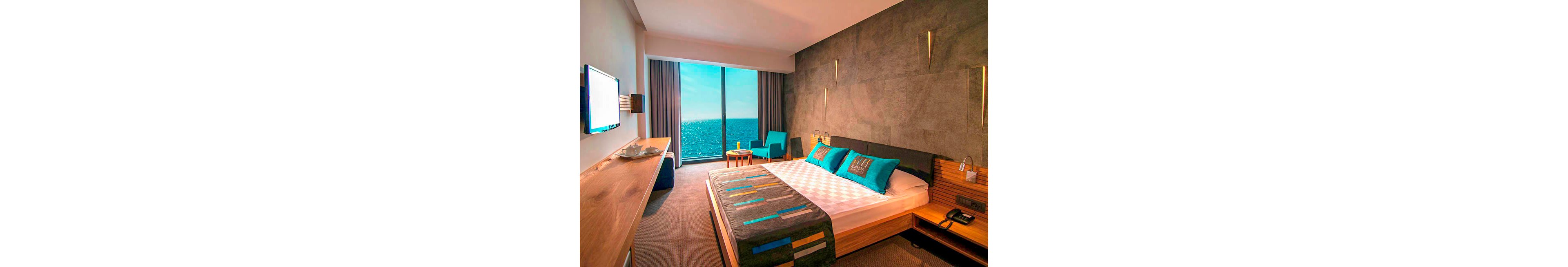 A contemporary hotel bedroom features a large bed with turquoise pillows and a patterned runner. Floor-to-ceiling windows open onto a bright ocean view, filling the room with natural light. Warm wooden furniture, including a desk and bedside tables, contrasts with the gray accent wall. Subtle wall lighting and minimalist decor create a calm, vacation-ready atmosphere.